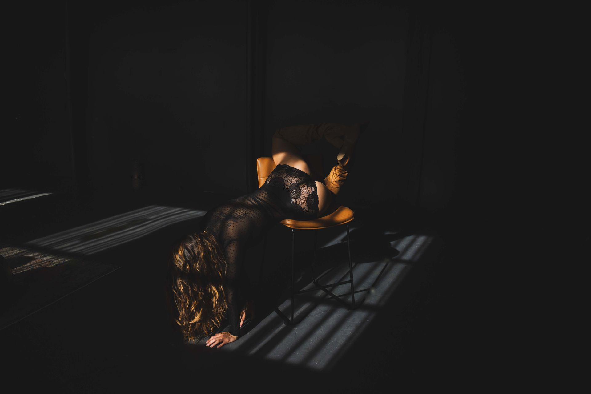 Woman in black lace leans over a chair, head down, lit by sunlight through blinds in a dark room.
