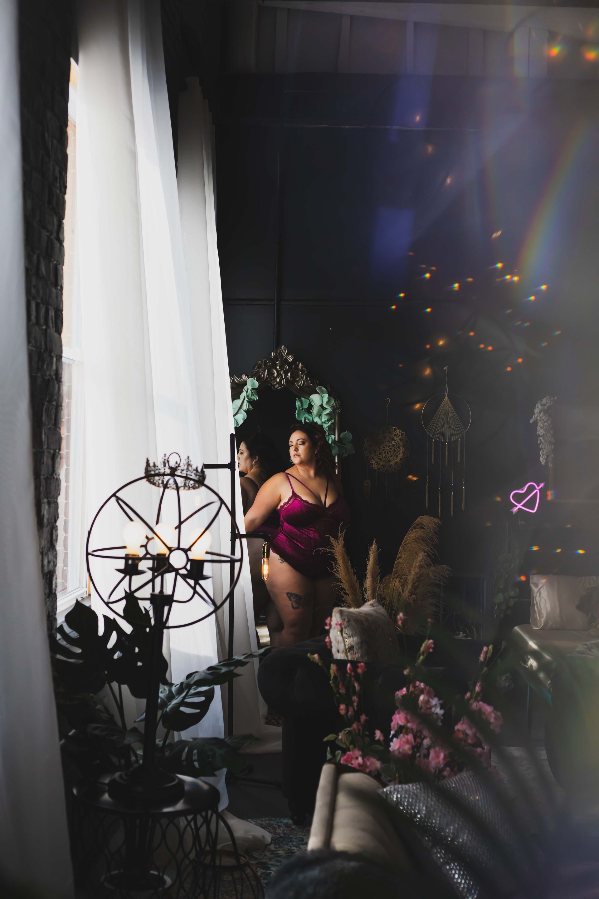 Woman in a magenta negligee, standing by a window, reflected in a mirror, amidst ornate decor.