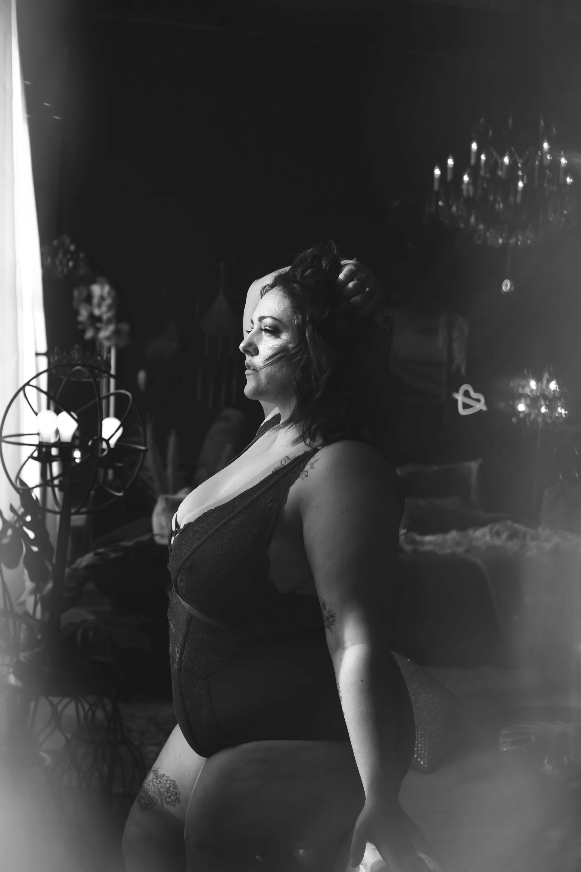 A woman in black lingerie poses in a dimly lit room, hand in her hair, gazing to the side.  A chandelier and other decor are visible.