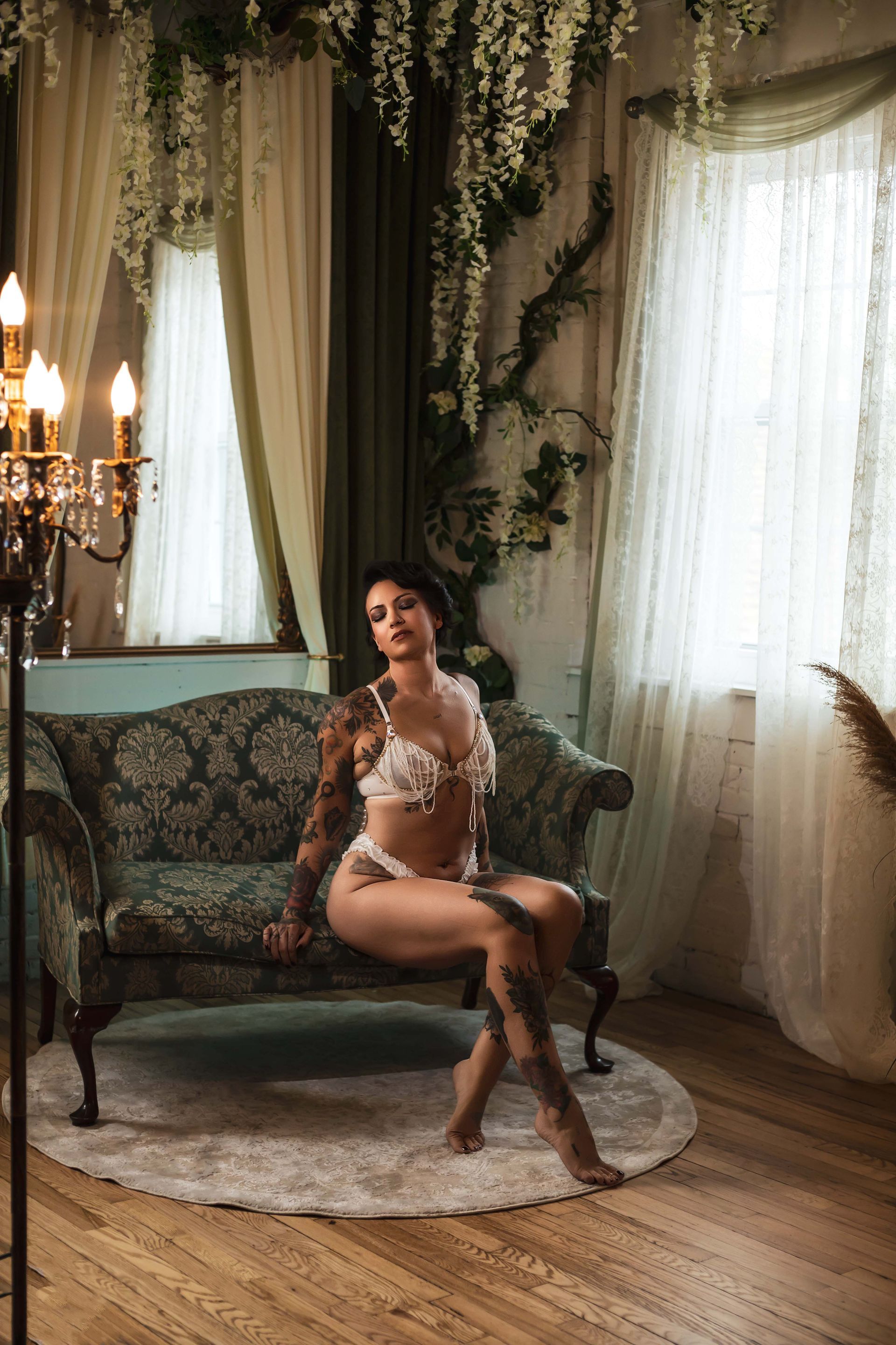 Woman in white lingerie sits on a green velvet sofa in a room with floral decor and a window. She has tattoos and a relaxed expression.
