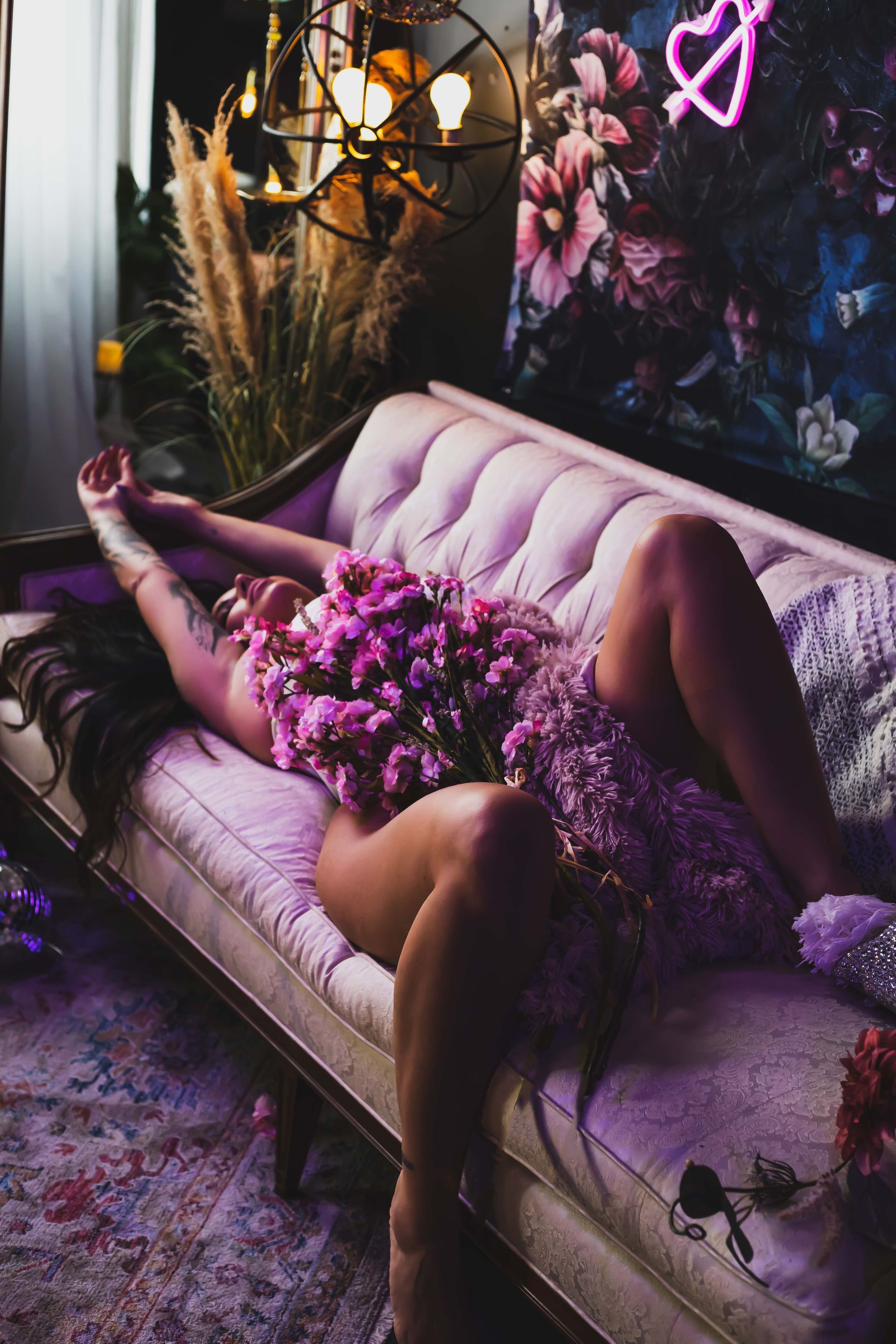 Woman reclines on a white couch, body partially covered with purple flowers. Dark room with floral art and pink neon heart.
