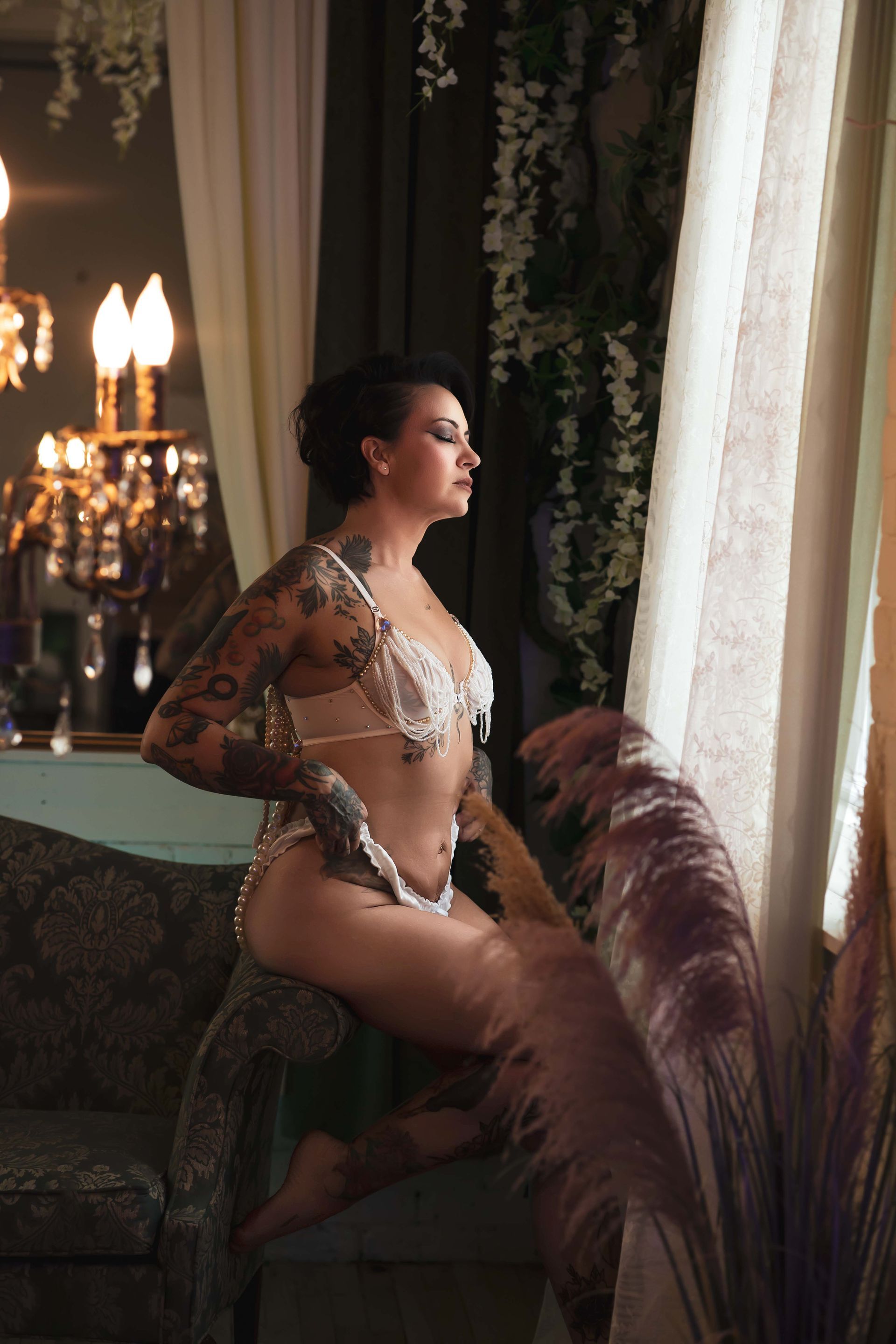 Woman with tattoos in white lingerie sits on a couch, looking out a window in a luxurious setting.