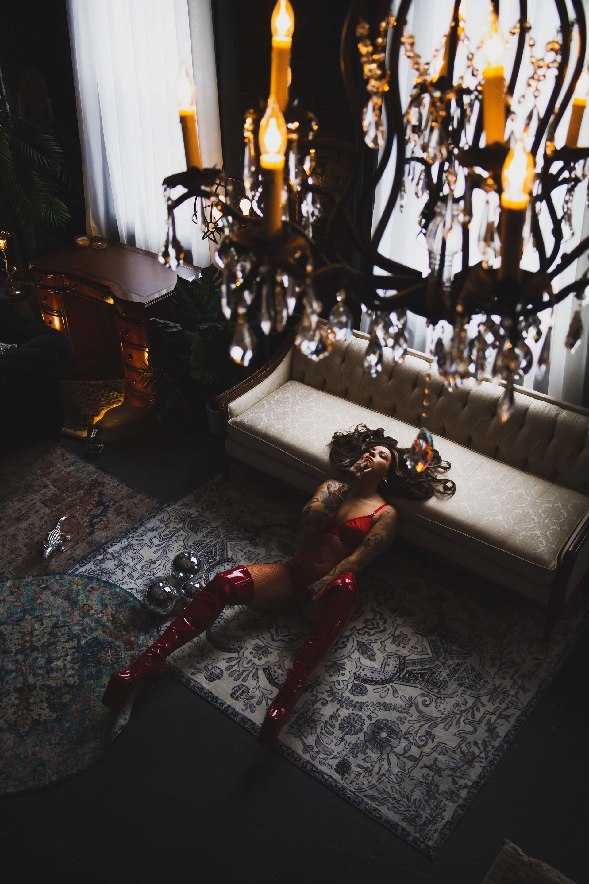 Woman in red lingerie and boots reclines on a patterned rug near a sofa, beneath a lit chandelier.