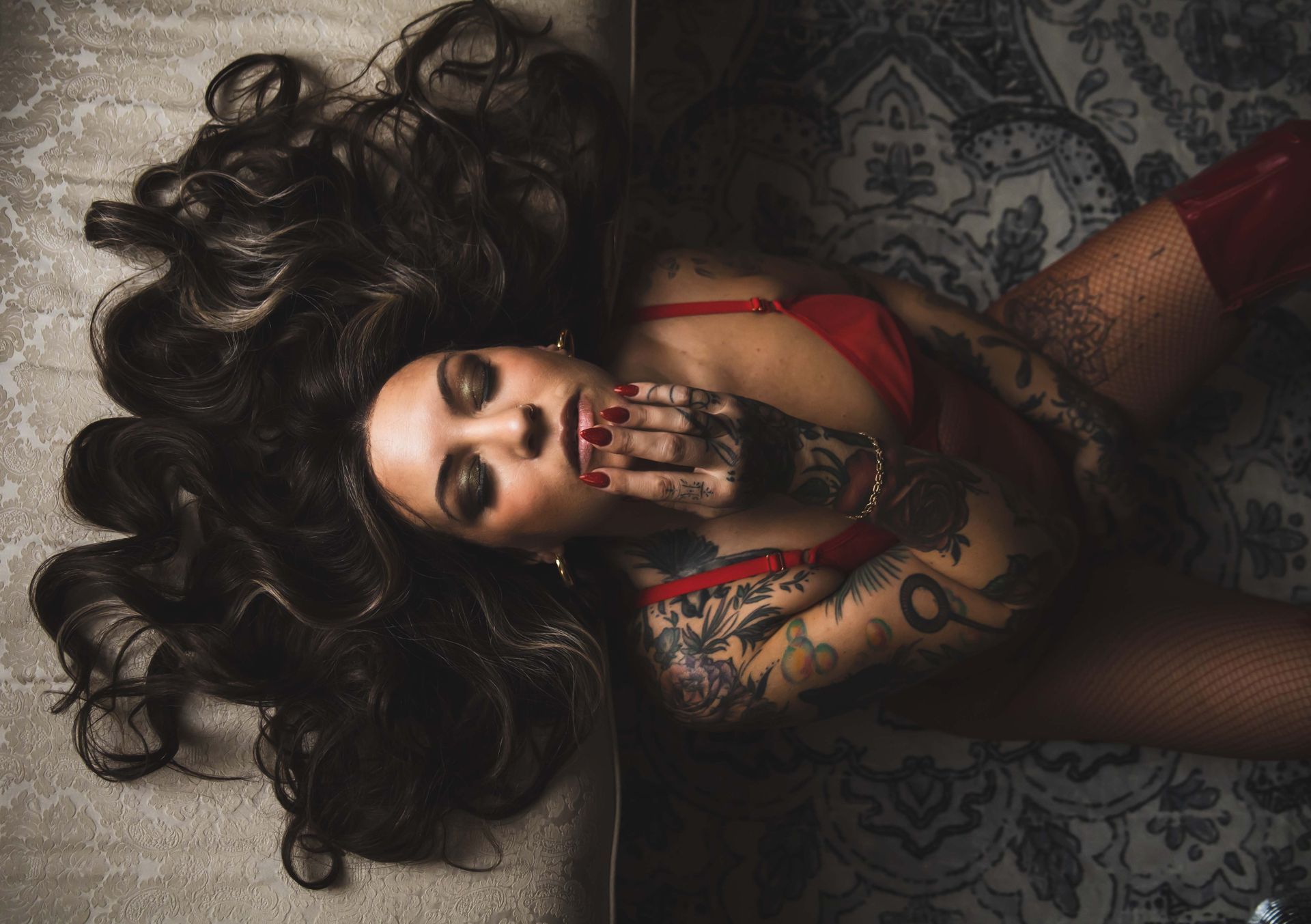 Woman with long dark hair and tattoos, wearing red lingerie and boots, lying on a patterned surface, covering her mouth.