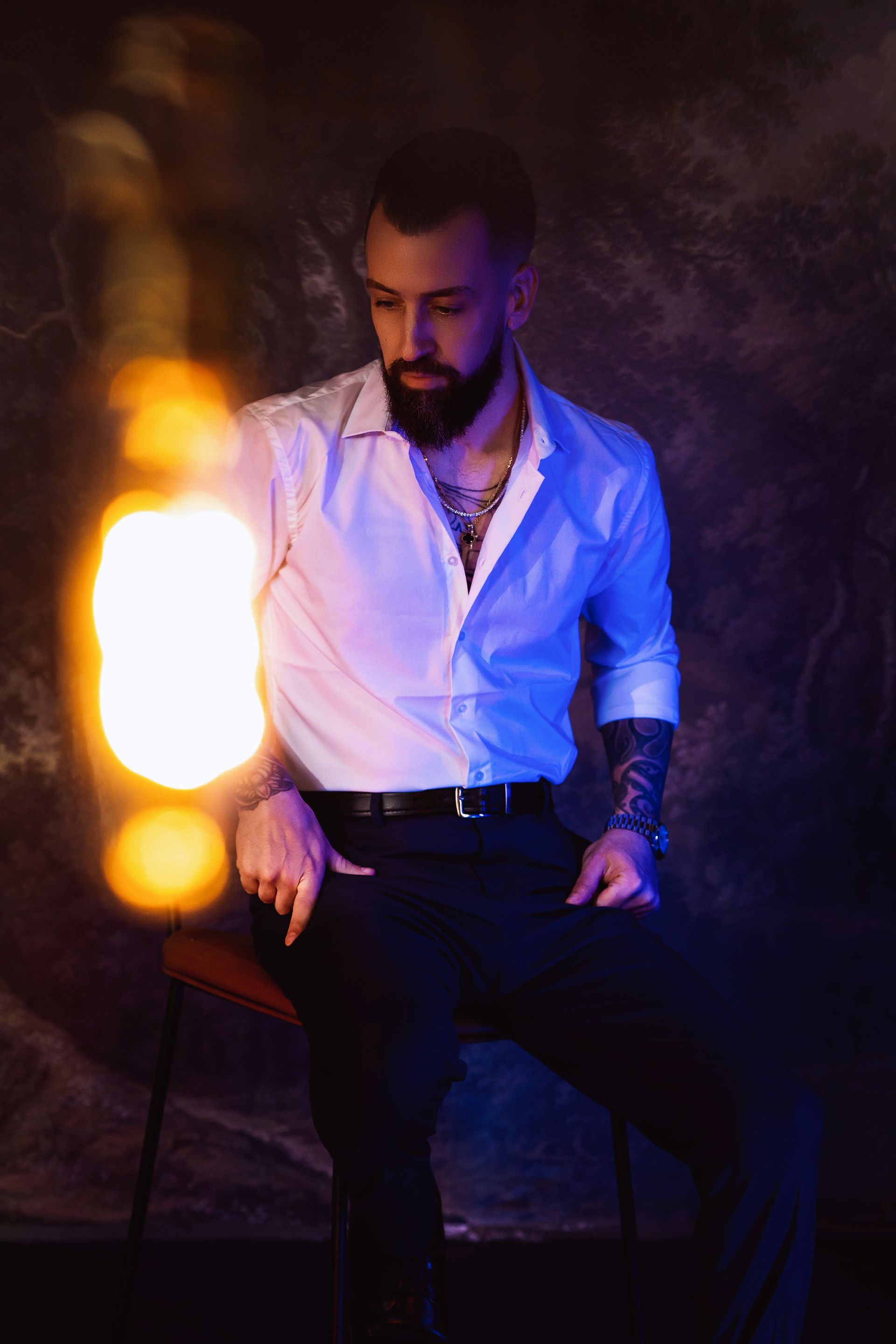 Man with beard and tattoos holds a glowing lantern, seated, wearing a white shirt and black pants.