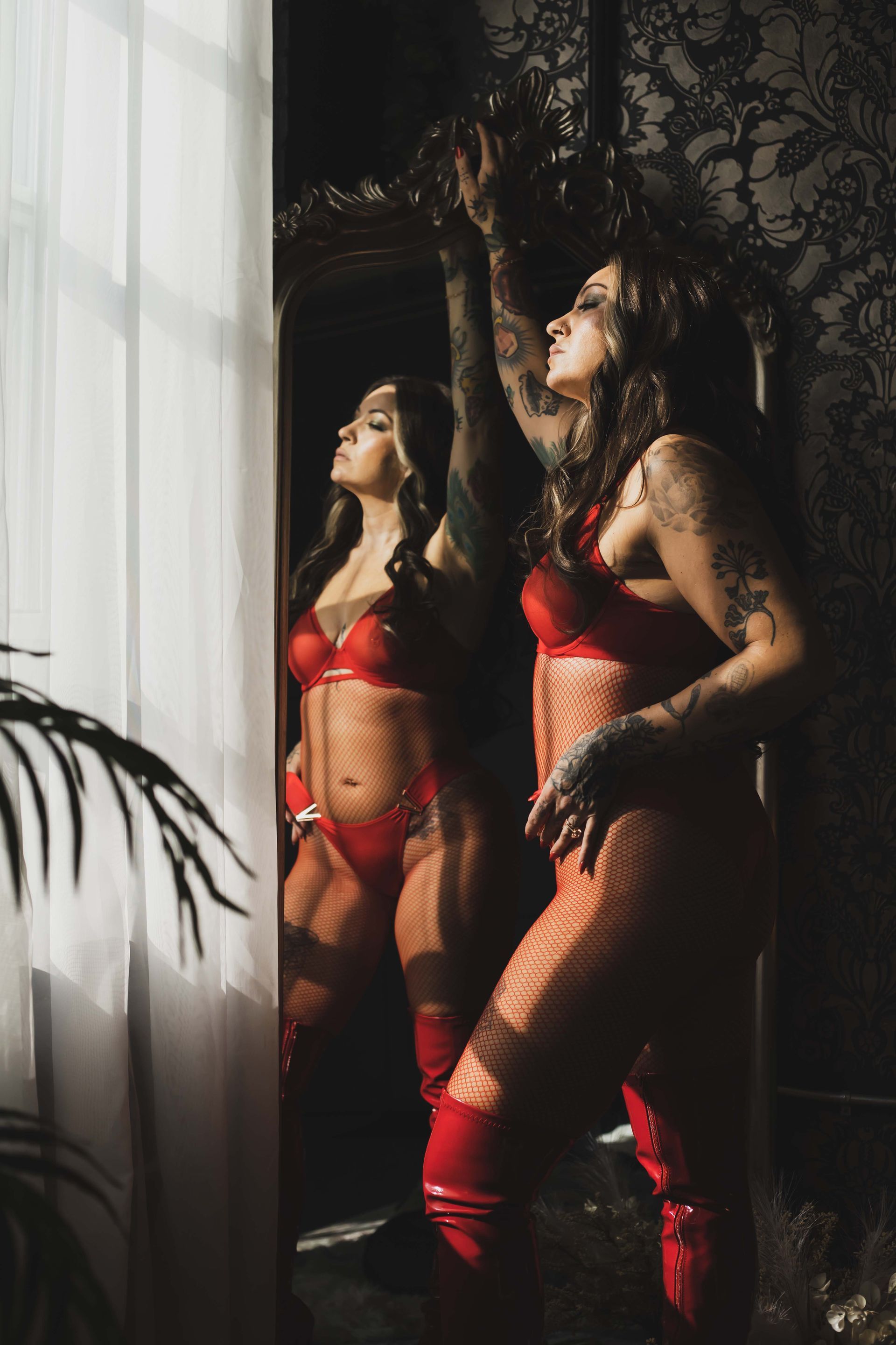 Woman in red lingerie poses by a mirror. She has tattoos and is reaching up towards sunlight from a window.