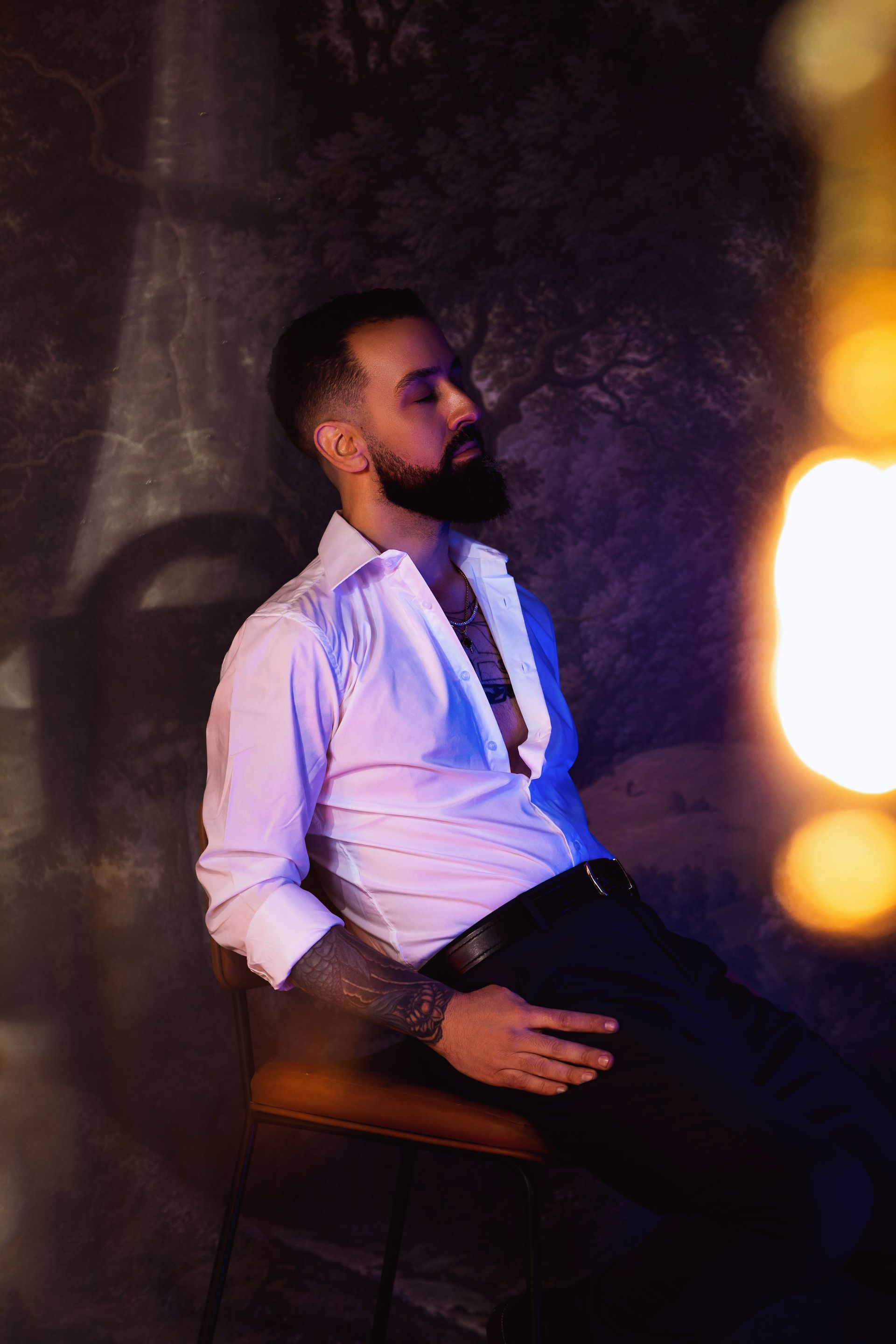 Man with a beard, in a white shirt, sitting, eyes closed. Tattoo on his arm, dark background, lit with blue and orange light.