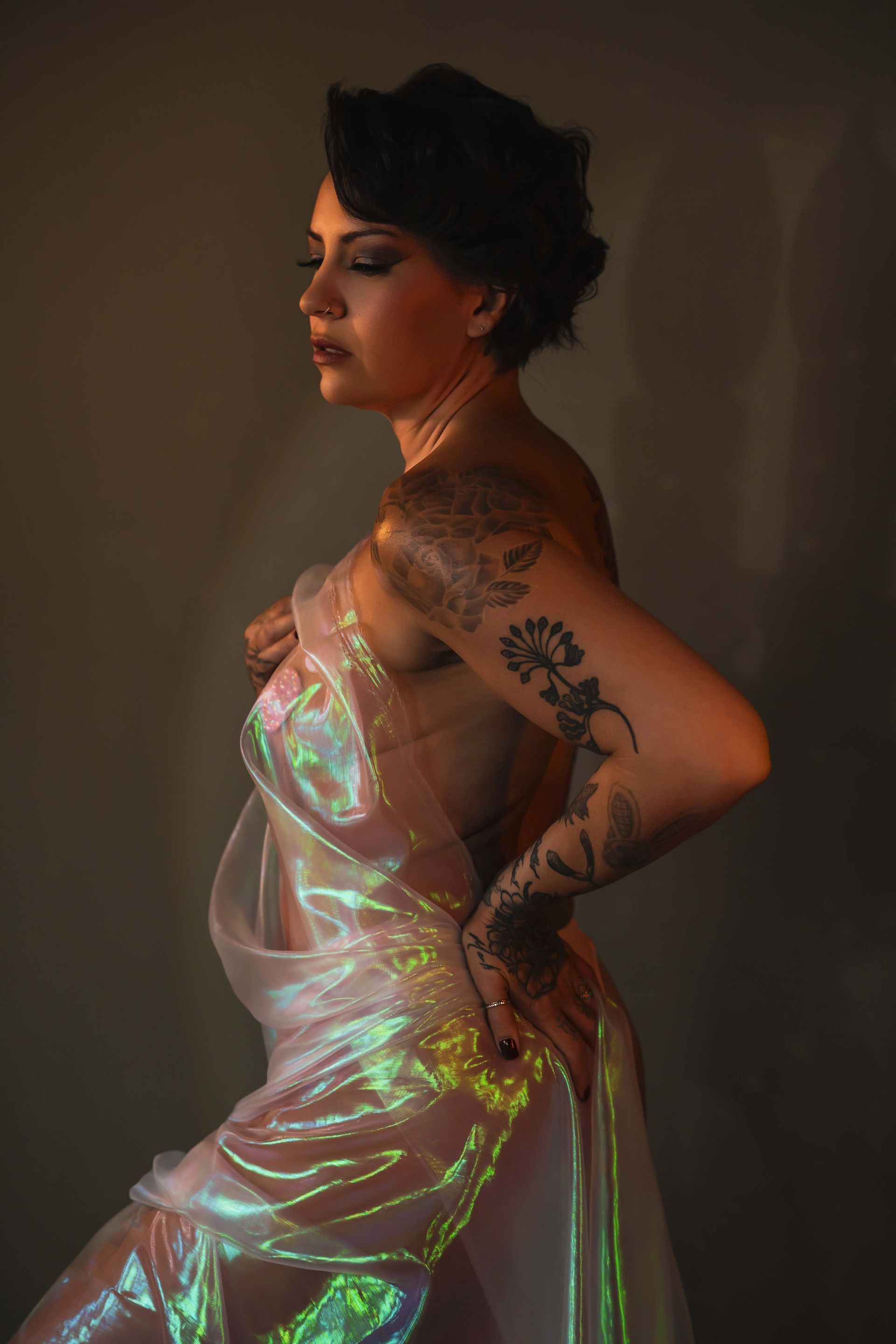 A woman with dark hair and tattoos draped in iridescent fabric, posing against a neutral background with warm lighting.