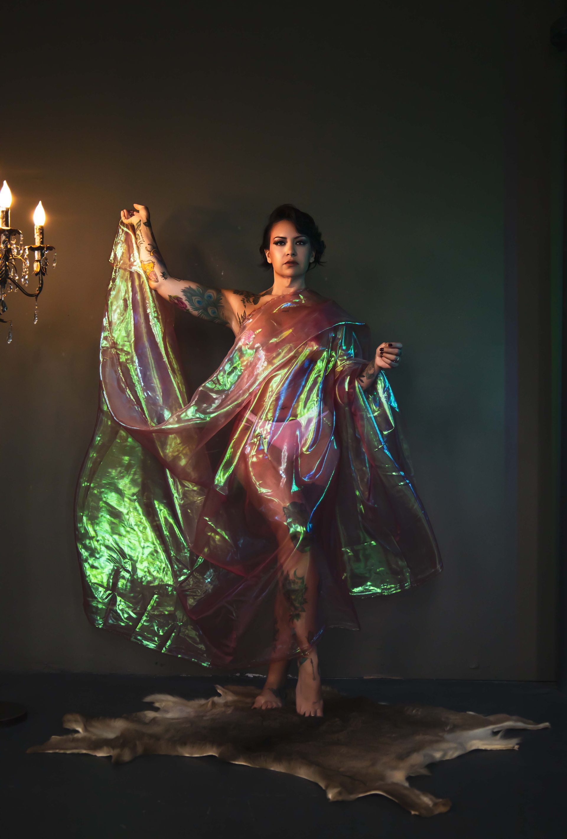 Woman in iridescent gown, posed dramatically in front of a dark wall, holding a flowing fabric. A candlelit chandelier is in the background.