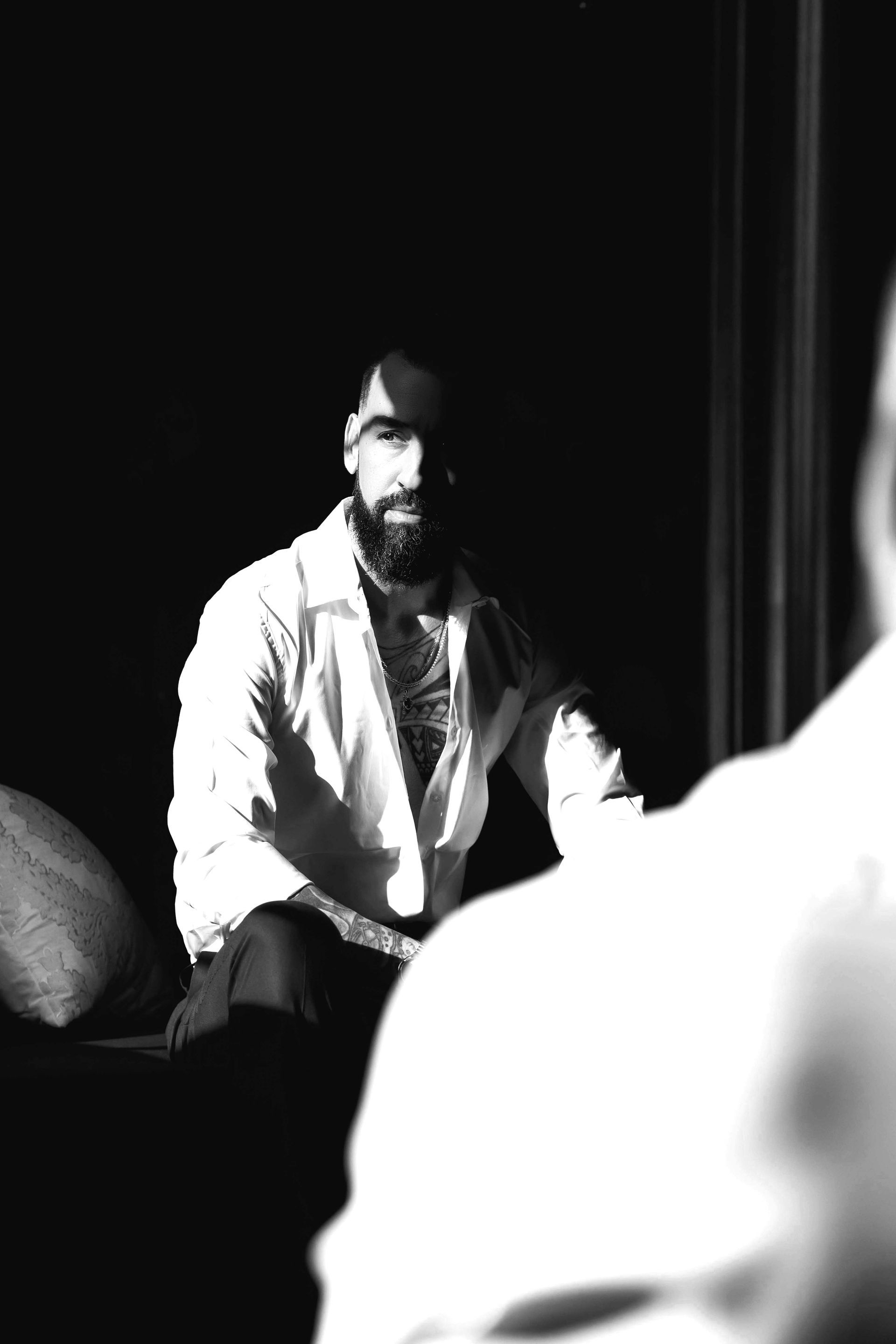 Man with a beard in a white shirt, seated and looking towards the camera, in a darkened room.