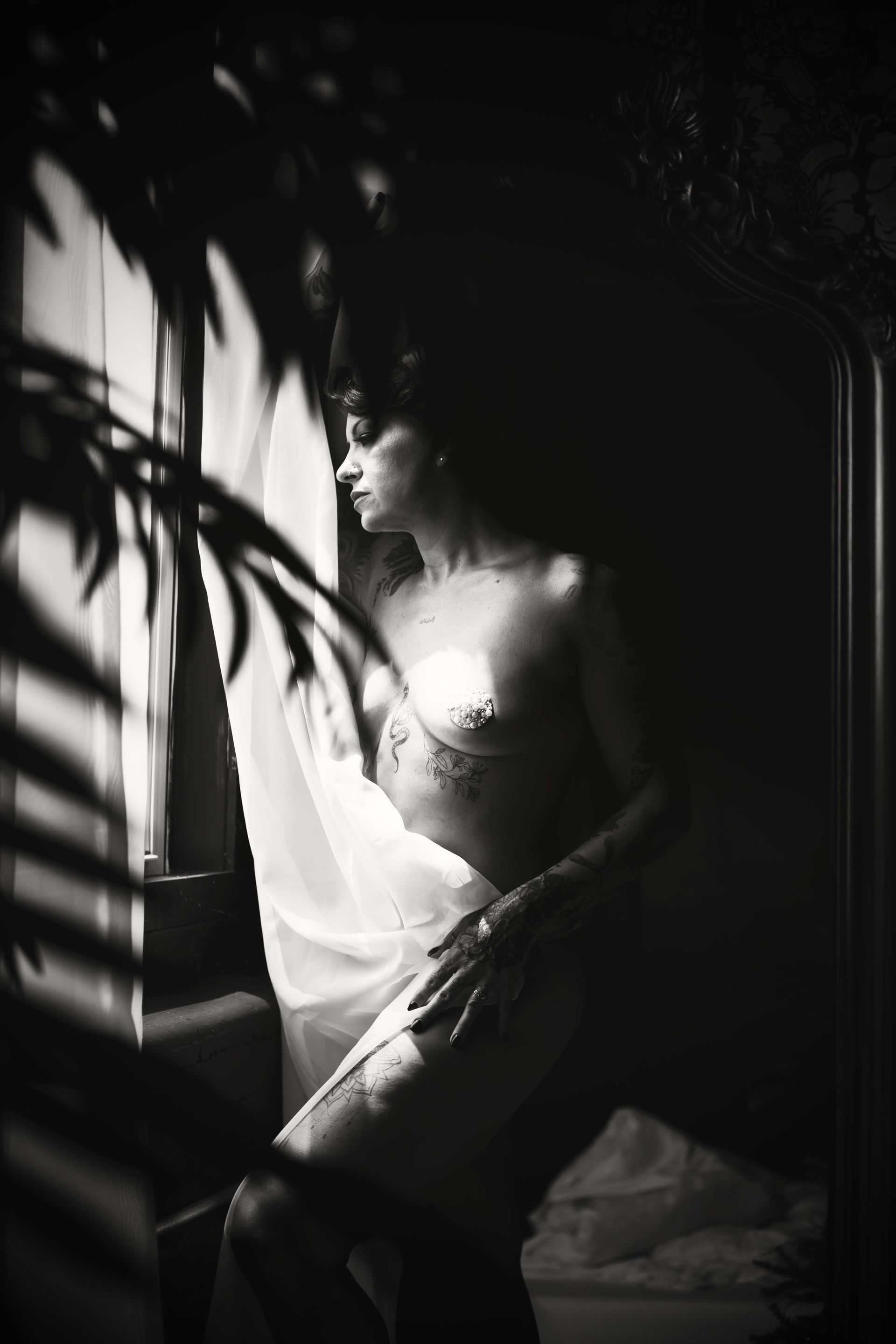 Black and white photo of a person standing near a window, partially covered by sheer fabric. Shadows from foliage fall across the scene.