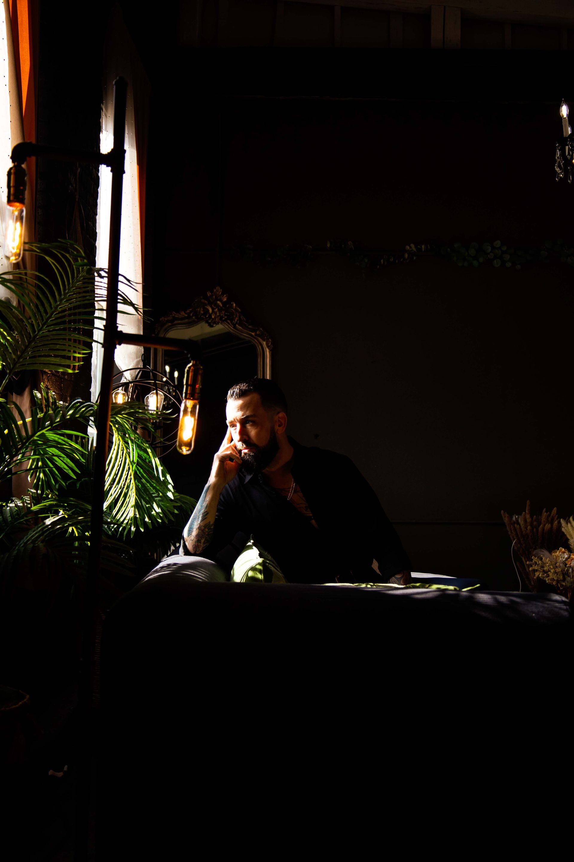 Man resting chin on hand, looking contemplative. Sitting at a table indoors. Dark lighting with visible plants and decorative bulbs.