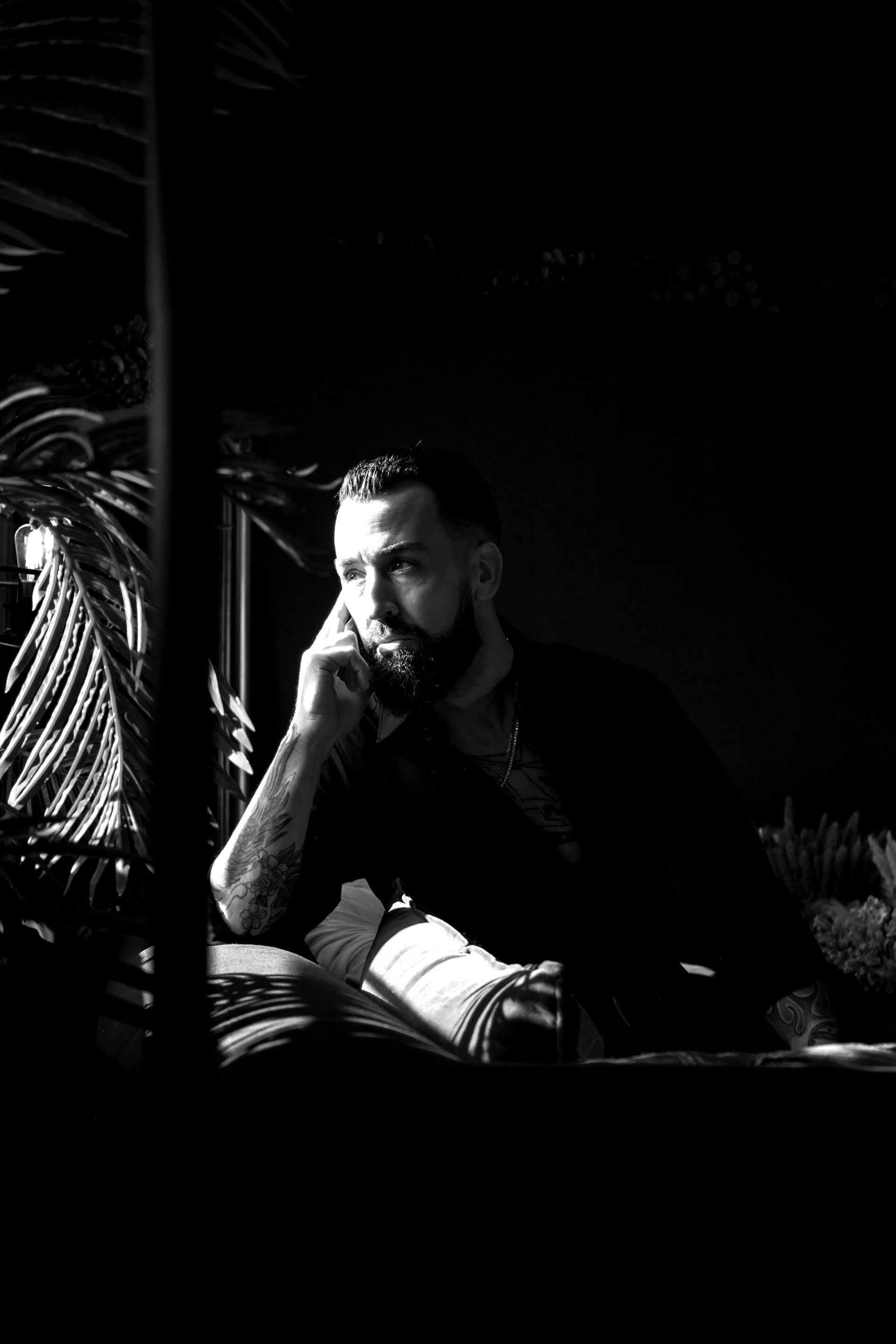 Man with beard resting chin on hand, lit by sunlight in dark setting.
