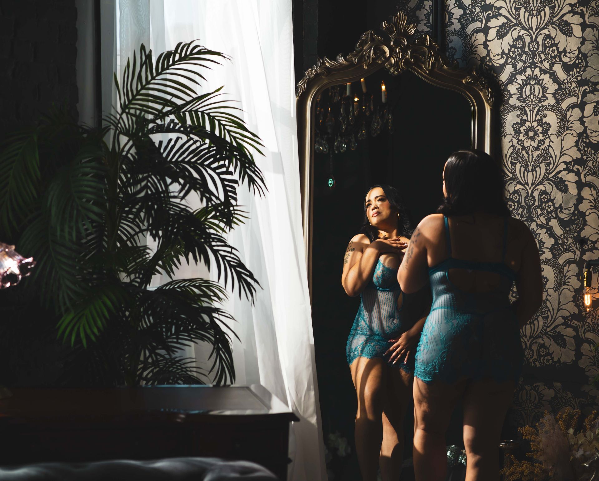 A woman in teal lingerie looks at herself in an ornate mirror. The room is dimly lit with a dark floral wallpaper and a large plant.