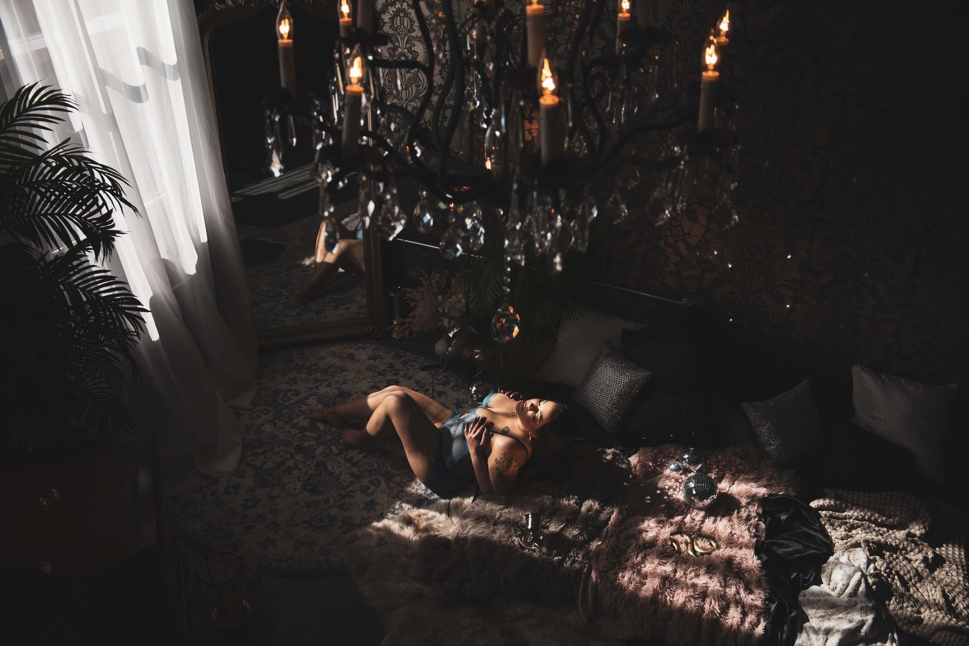 Woman reclines on a patterned rug in a dimly lit room, under a chandelier with candles.