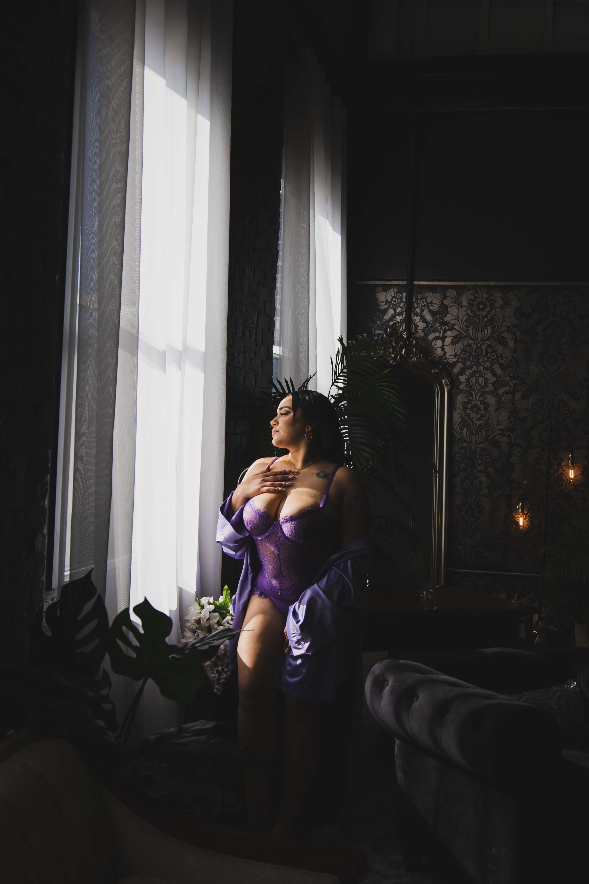 Woman in purple lingerie poses by a sunlit window. She looks out, hand on chest, in a moody, dark room.