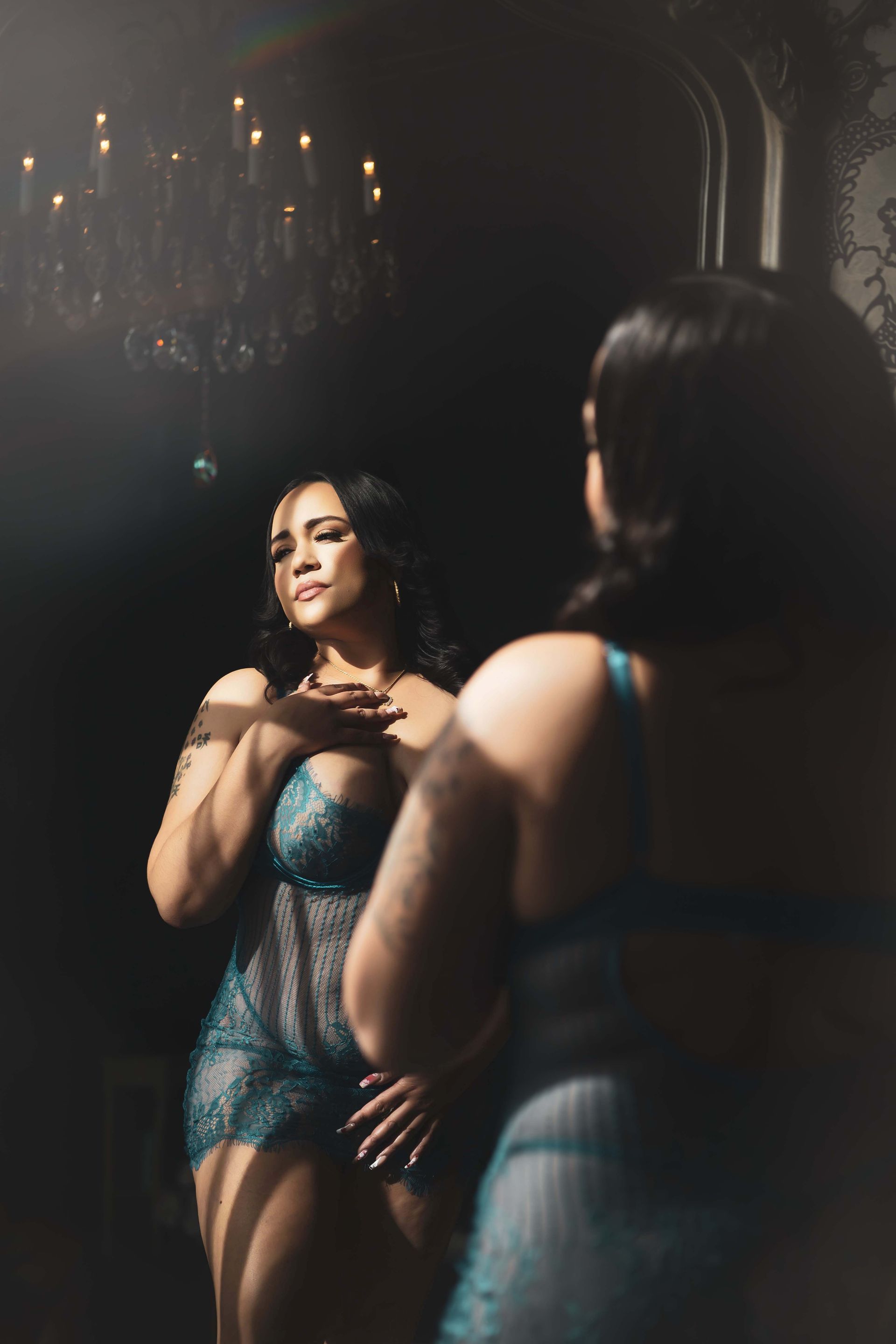 Woman in teal lingerie poses in front of a mirror, lit by a chandelier and sunlight. She touches her chest, eyes closed.