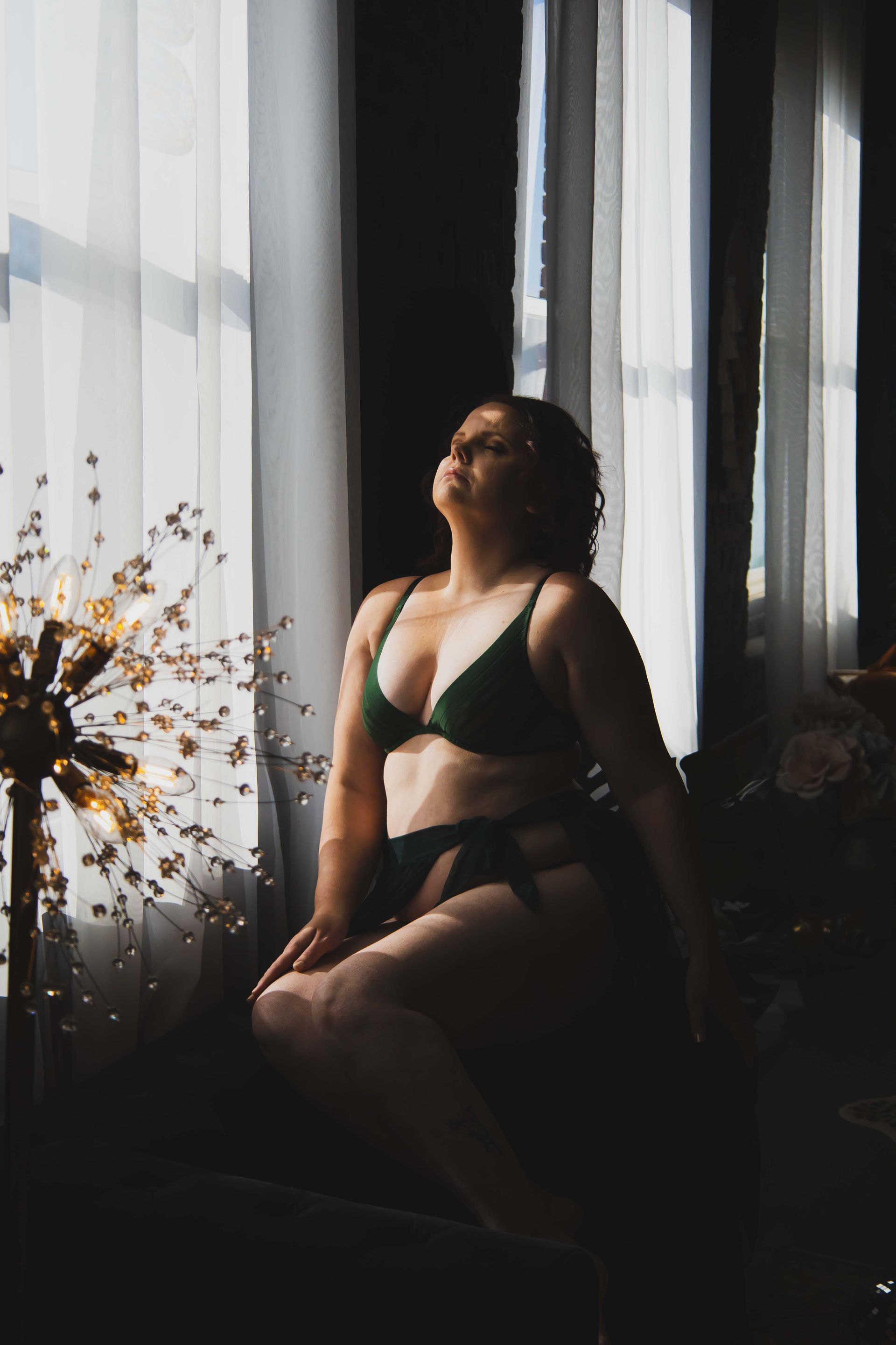 Woman in green lingerie sits near a window, bathed in sunlight. She looks upward with a serene expression in a dark, ornate room.