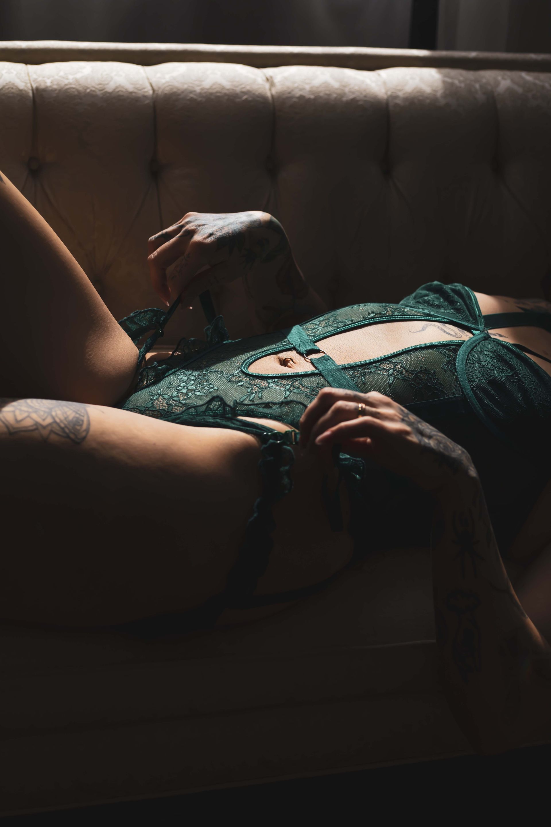 Woman reclines on a tufted couch, wearing a green lace bodysuit, hands near the garment.