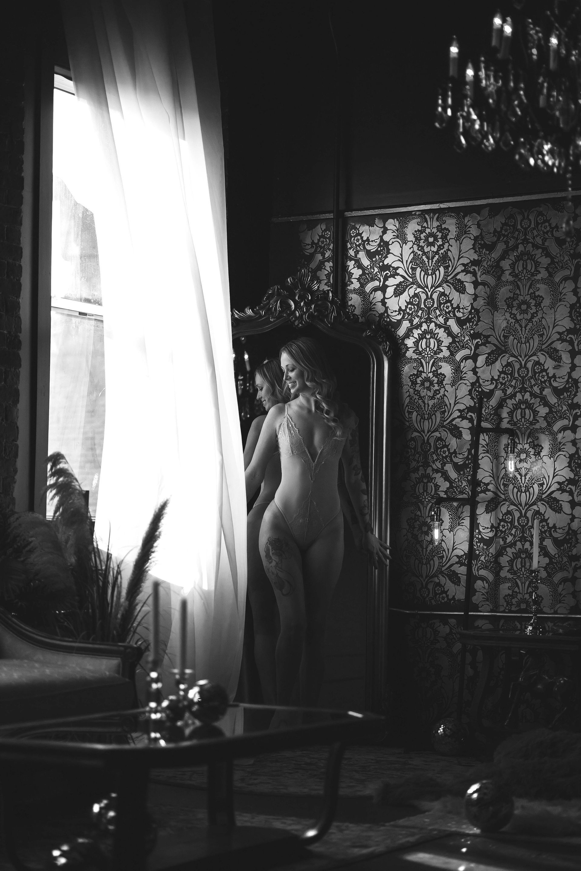 Black and white interior. A woman stands by a mirror, partially nude, looking out a window. The room has ornate furniture and a chandelier.