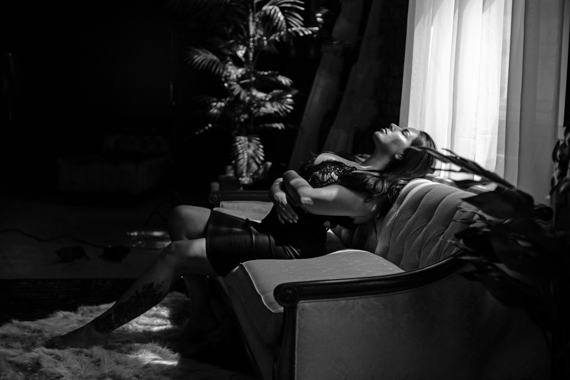 Woman reclining on a vintage couch in a dark room, lit by sunlight. Black and white with soft lighting.