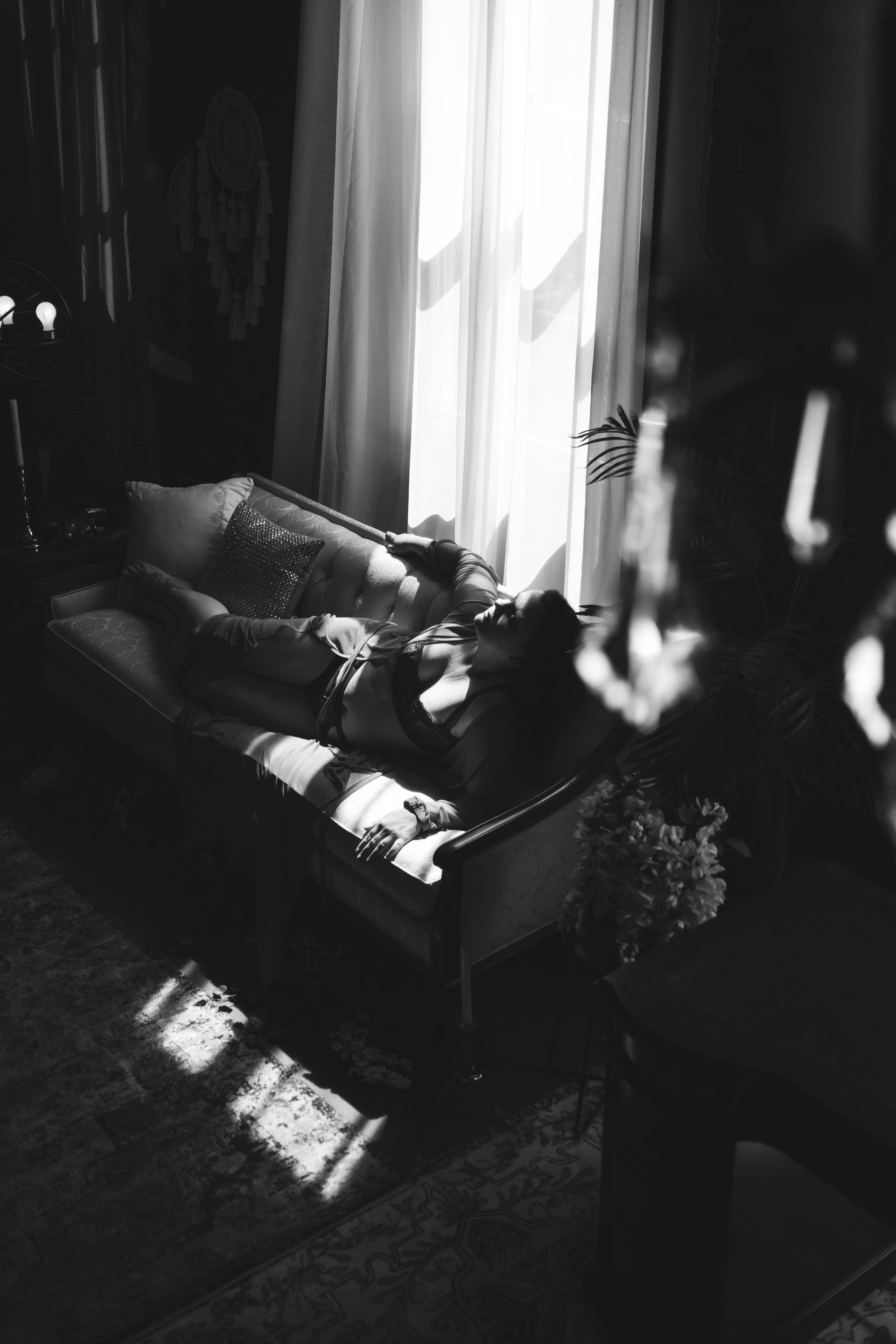 Woman reclines on a chaise lounge near a window, bathed in sunlight. Black and white photo, ornate setting.