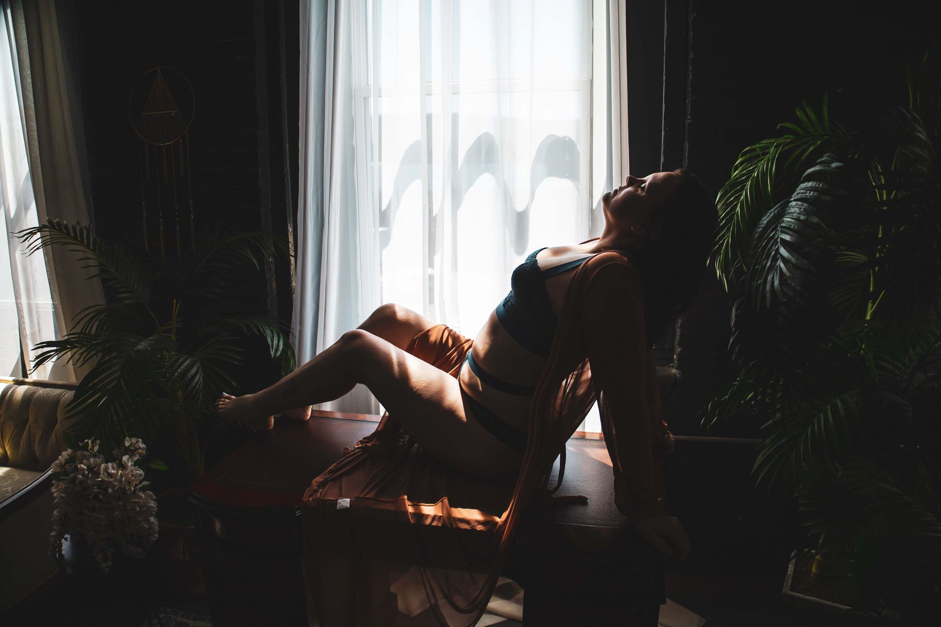 Woman in lingerie reclines on a dark surface, backlit by a window. She arches her back in a room with plants.