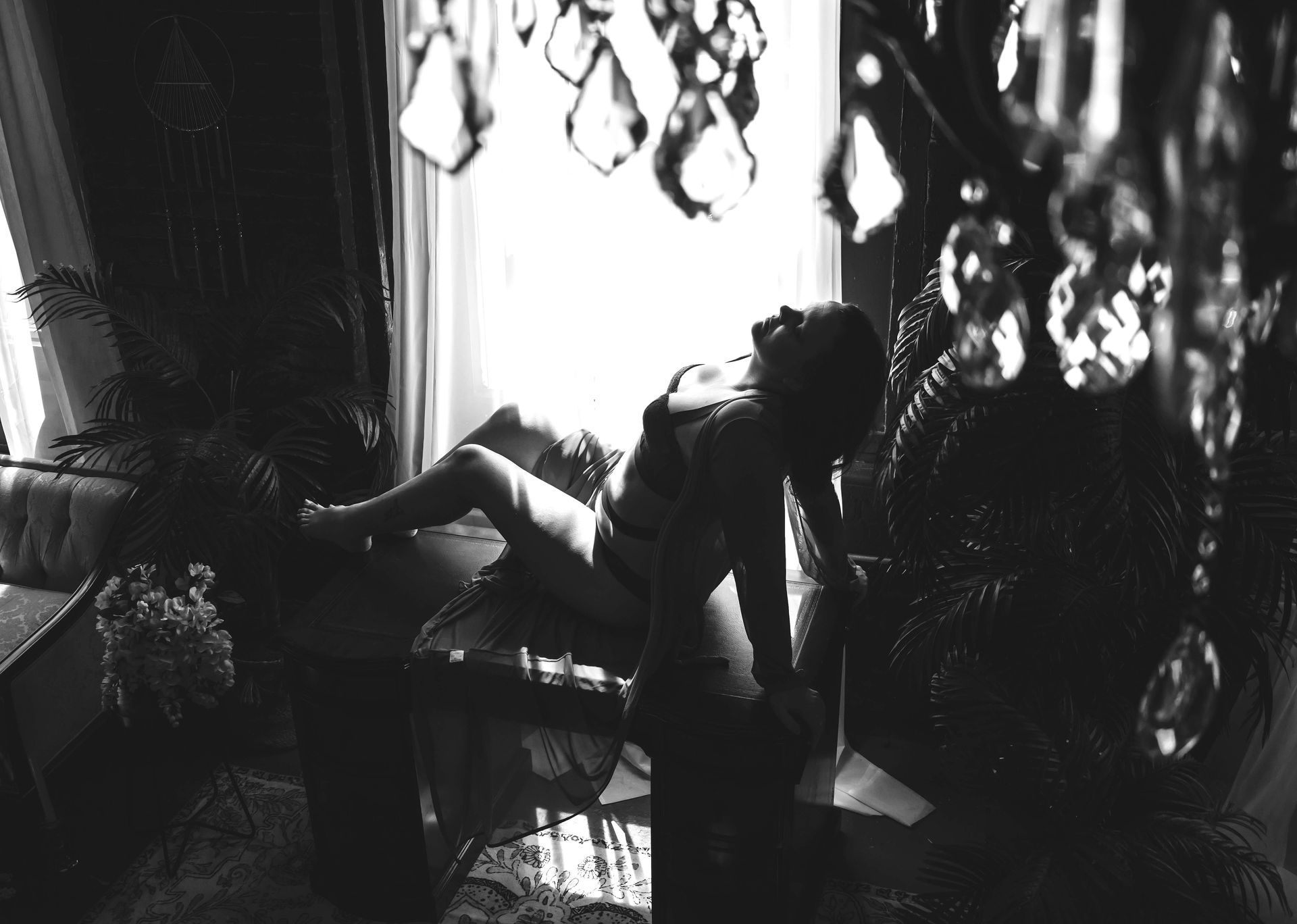 Black and white photo of a person on a bed in a room with a chandelier. Light from a window illuminates them.