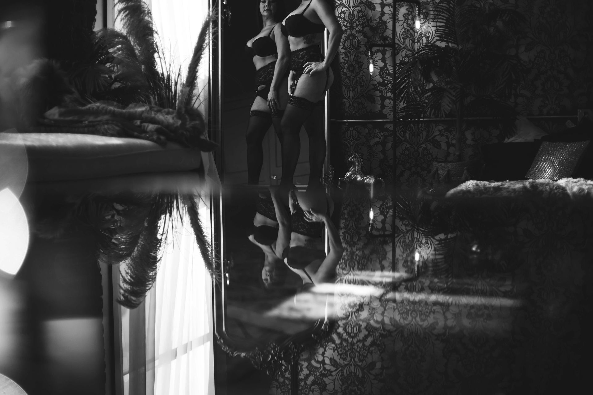 Woman in lingerie reflected in a mirror; bedroom setting with dim lighting and patterned wallpaper.
