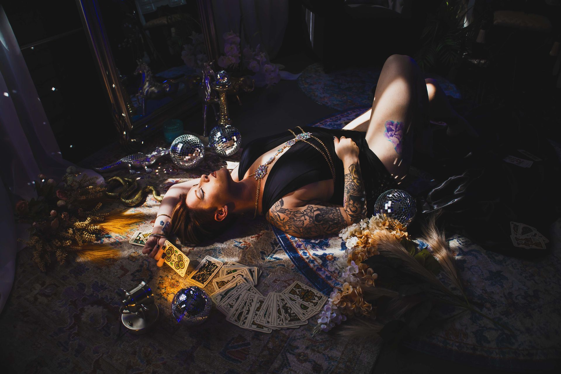 Woman with tattoos lies amongst tarot cards and decorations, bathed in light and shadow.