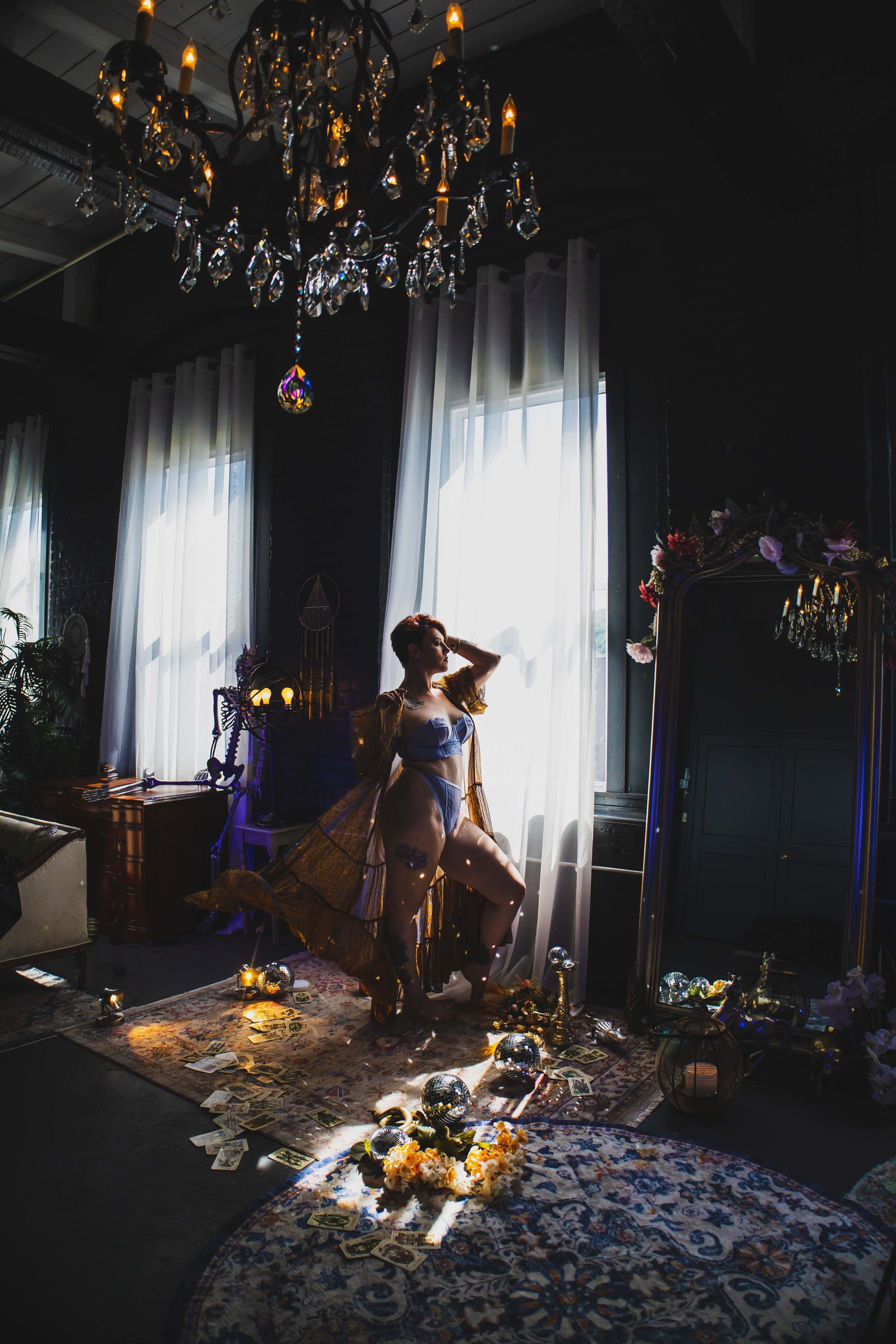 Woman in lingerie poses by a window, lit by sunlight. Elegant room with a chandelier and flowers.