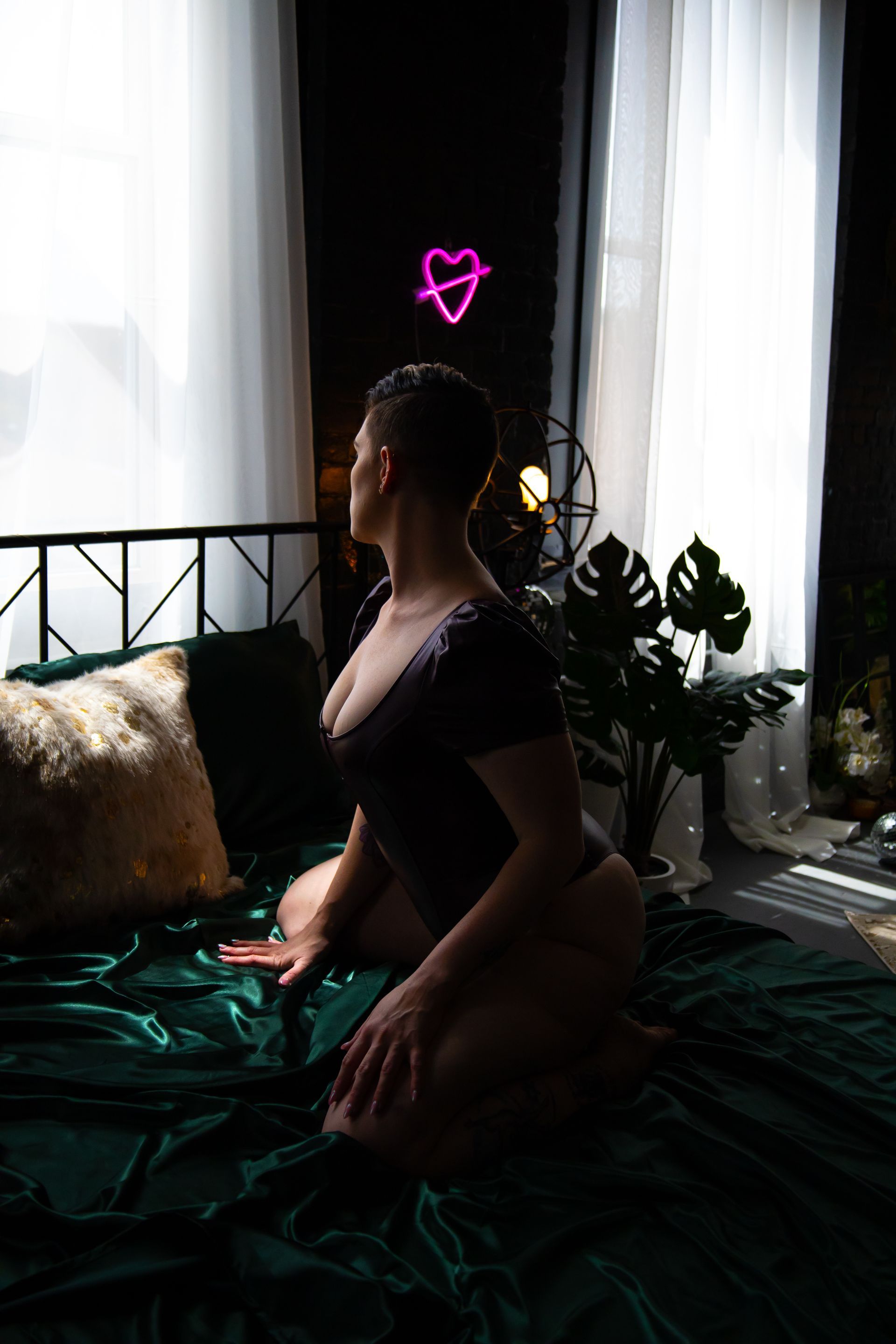 Woman with short hair kneels on a green velvet bed, looking towards a bright window. A neon heart illuminates the dark room.