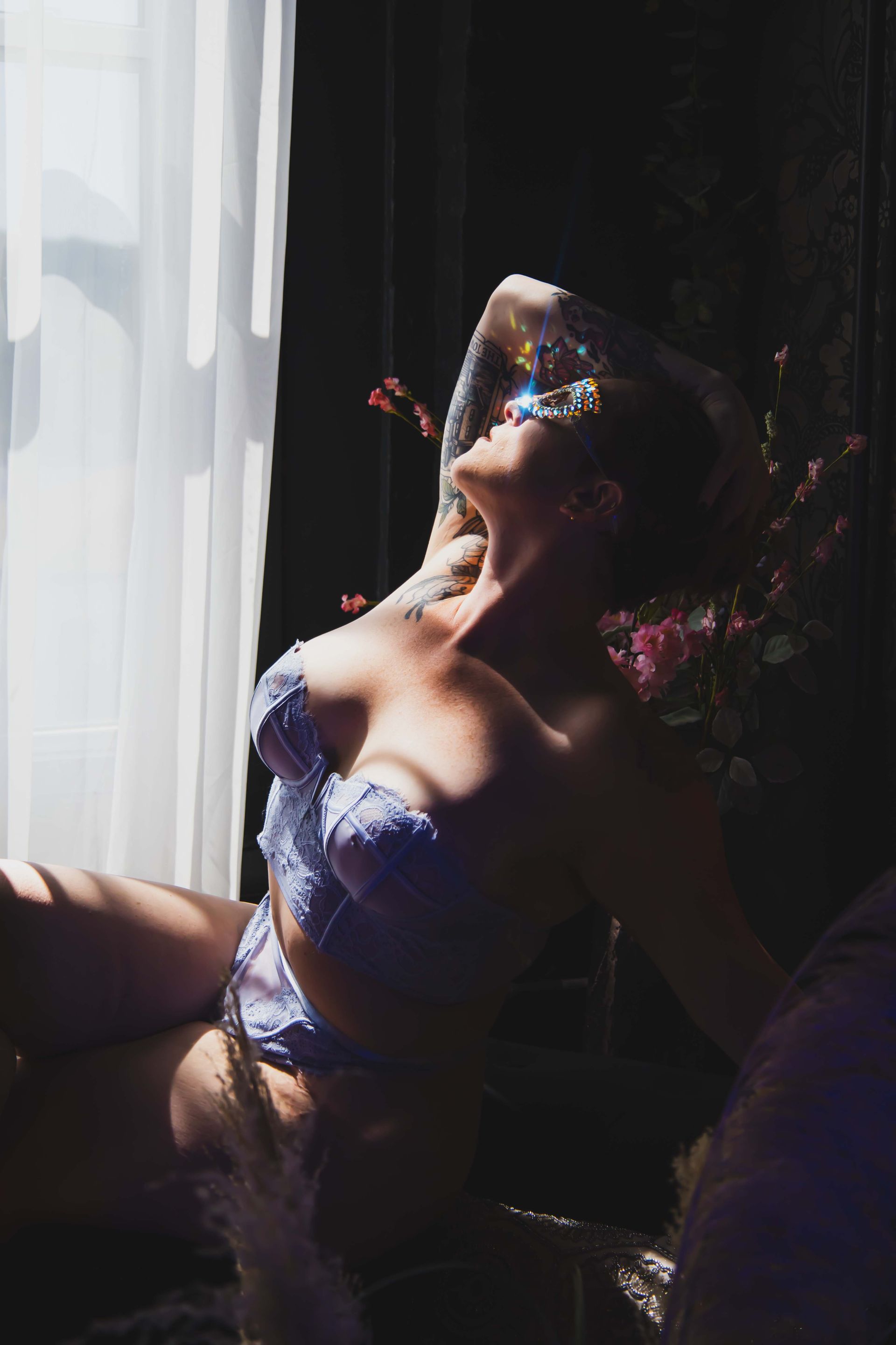 Woman in light purple lingerie reclines by a window, head tilted back, face partially obscured by sparkles. Dramatic lighting.
