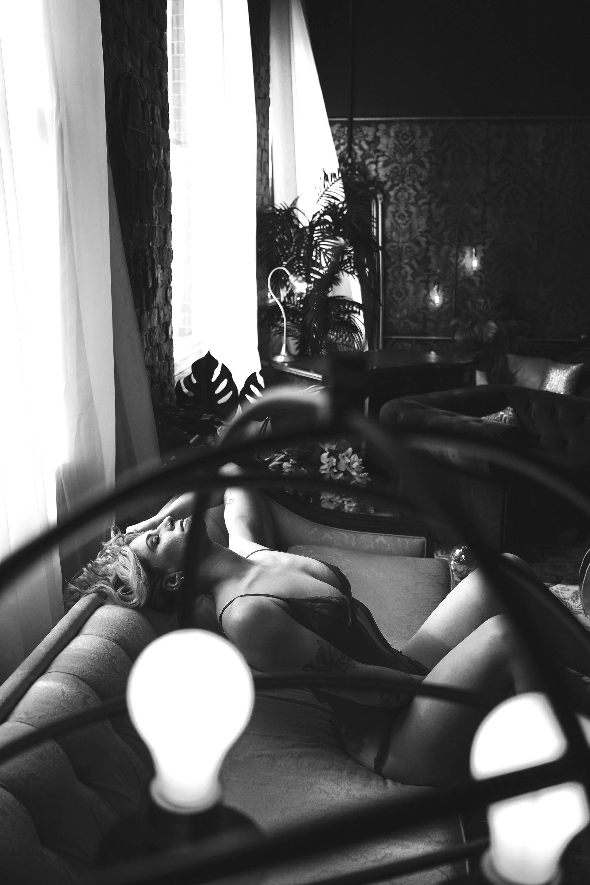 Woman reclines on a bed, lit by soft light, with a curtain in the background. Black and white.