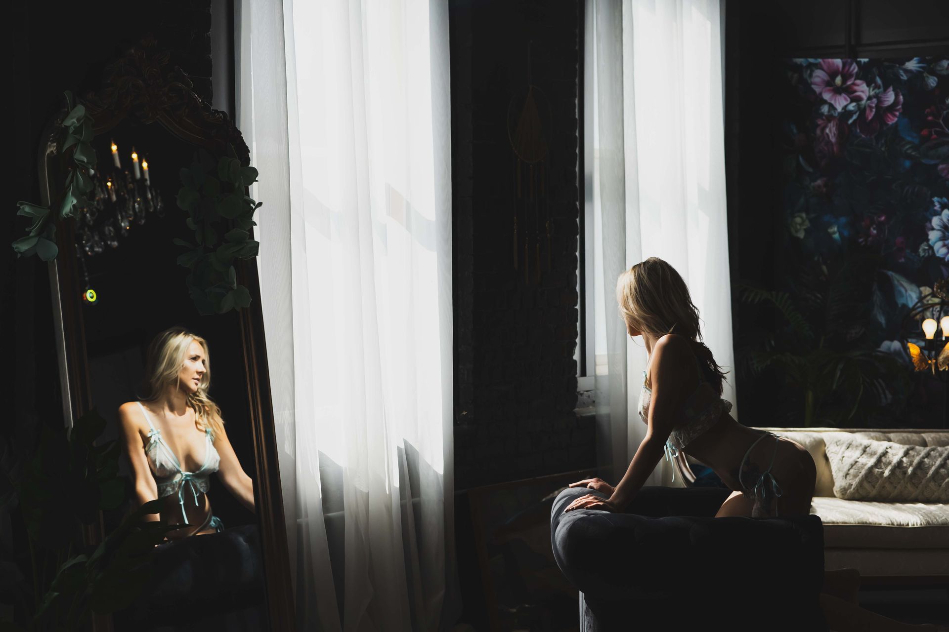 Woman in lingerie poses near a window, reflected in a mirror. Soft lighting and dark background create a sensual atmosphere.