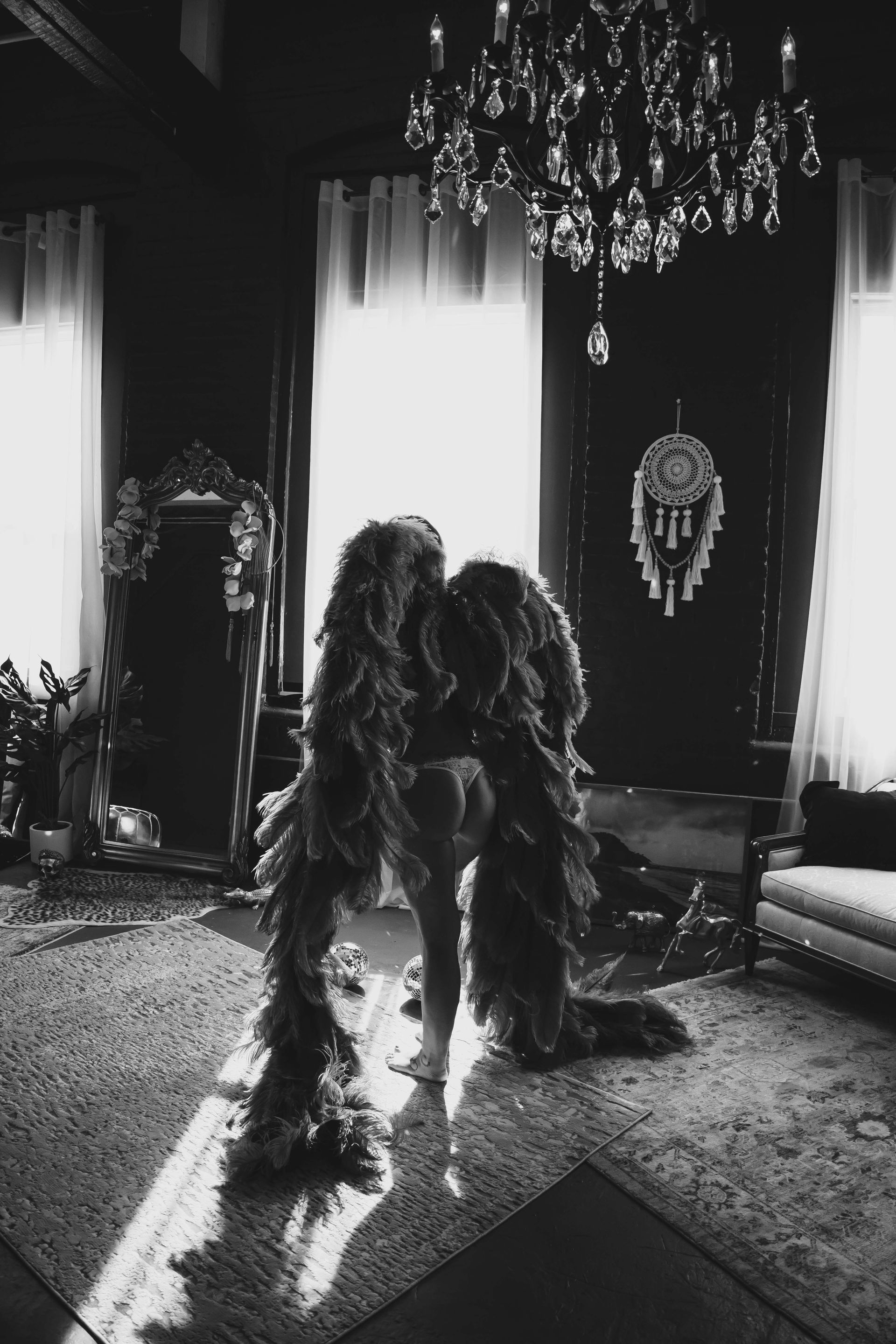 A person cloaked in long, feathery material stands in a dimly lit room with a chandelier and a dream catcher.