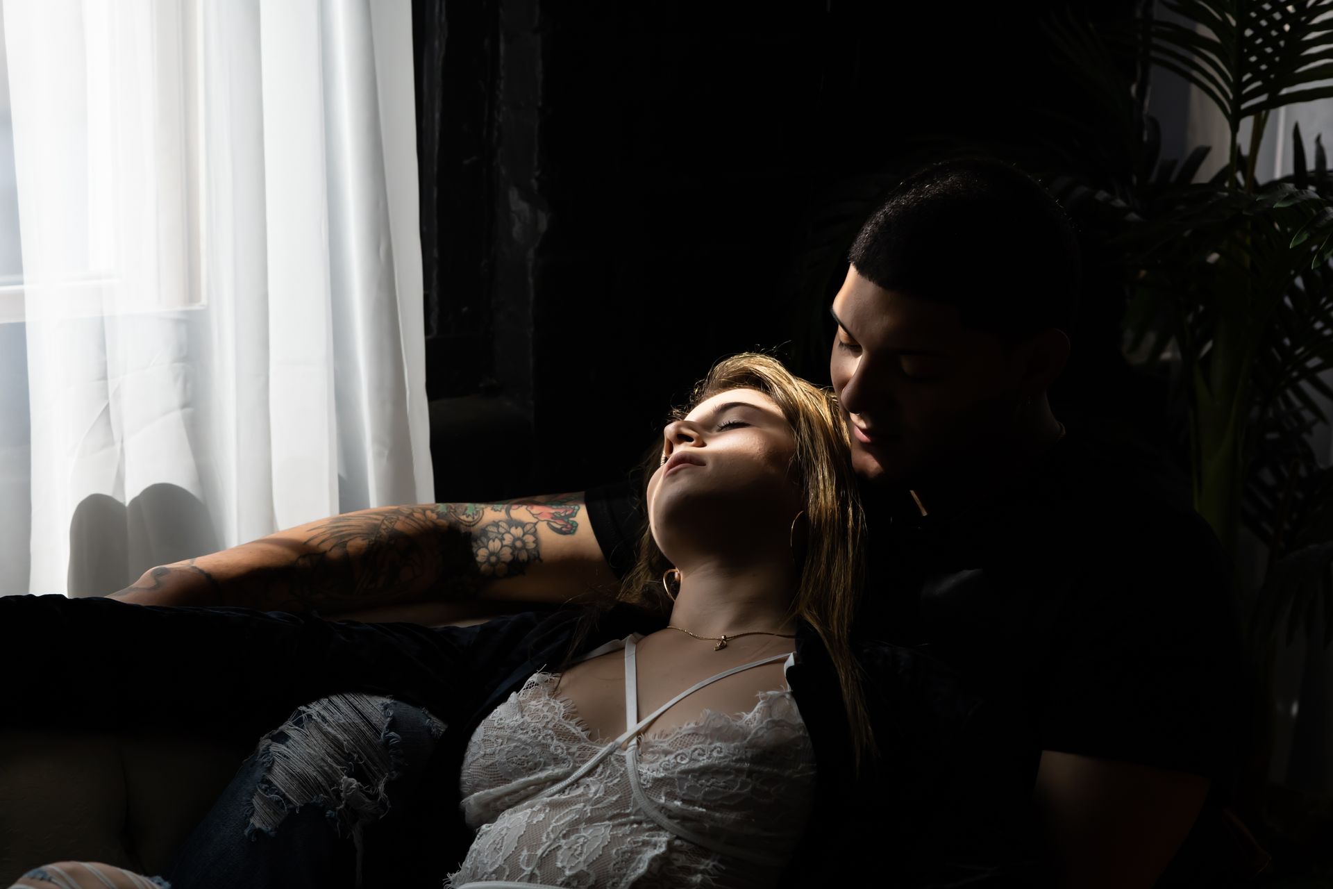 A couple embraces intimately on a sofa by a window. The woman reclines with her eyes closed; the man looks at her.