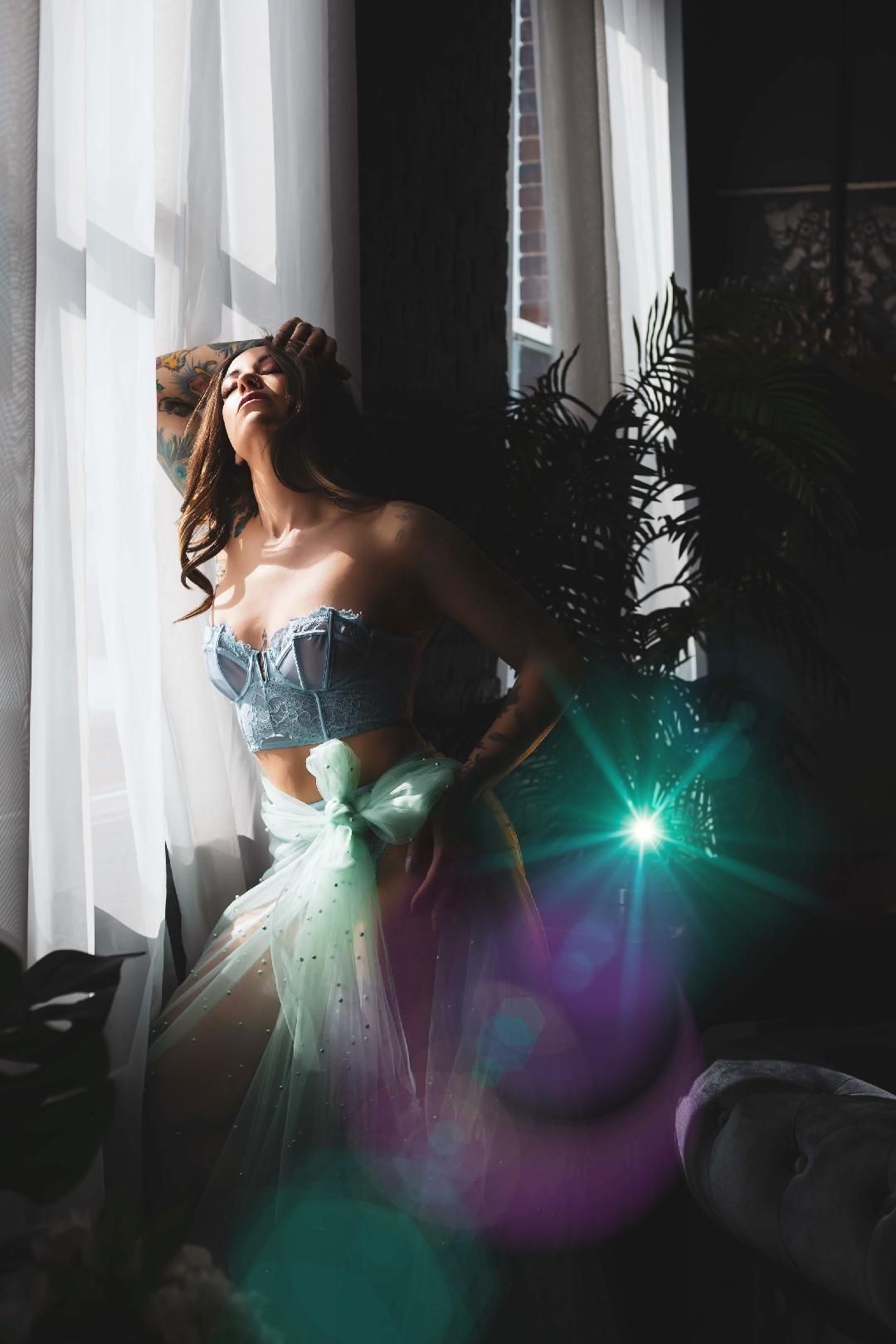 Woman in light blue corset and sheer skirt poses near a window, bathed in sunlight, with a green bow detail.