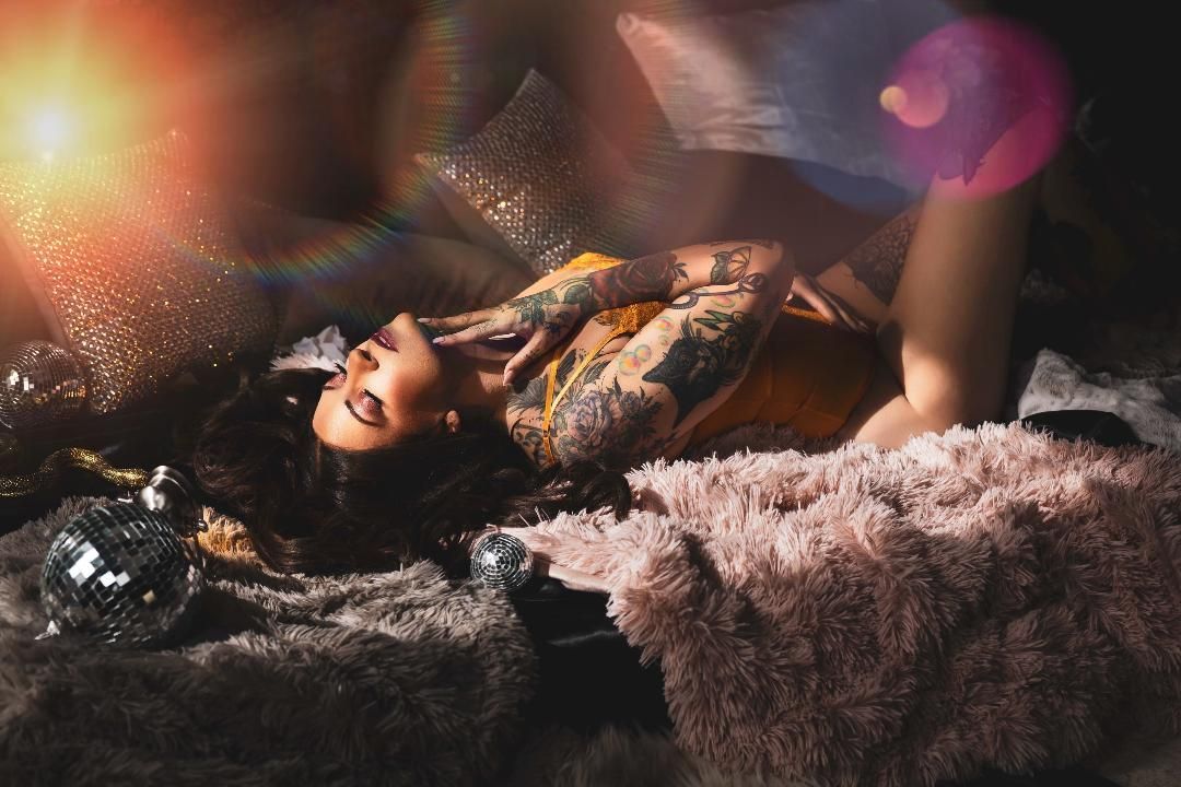 Woman with tattoos reclines on a plush gray blanket, looking upward. She wears a yellow top, and the setting is dimly lit with soft light.