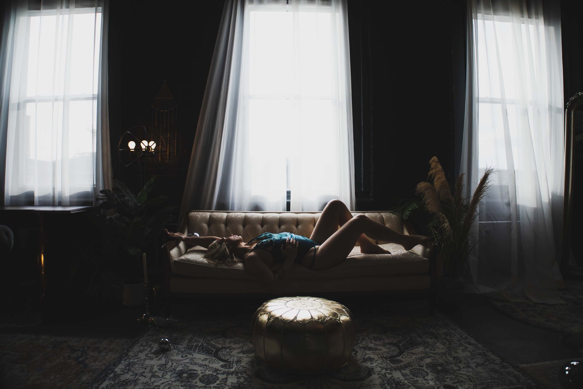 A woman lies on a couch in a dimly lit room, with sunlight streaming in from large windows.