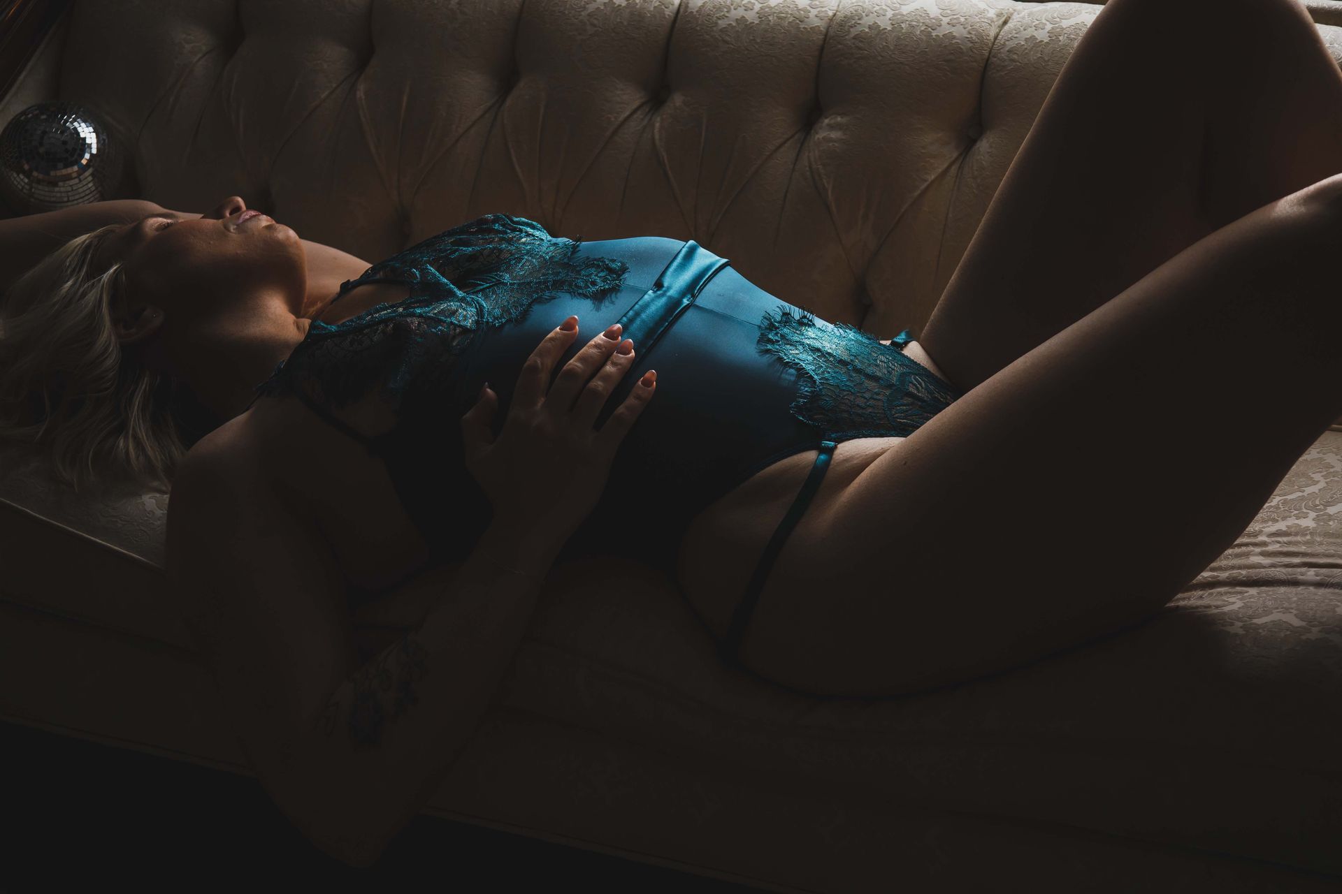 Woman in teal lingerie reclines on a tufted couch, illuminated by soft light. She lies with her legs open, her hands resting on her chest.