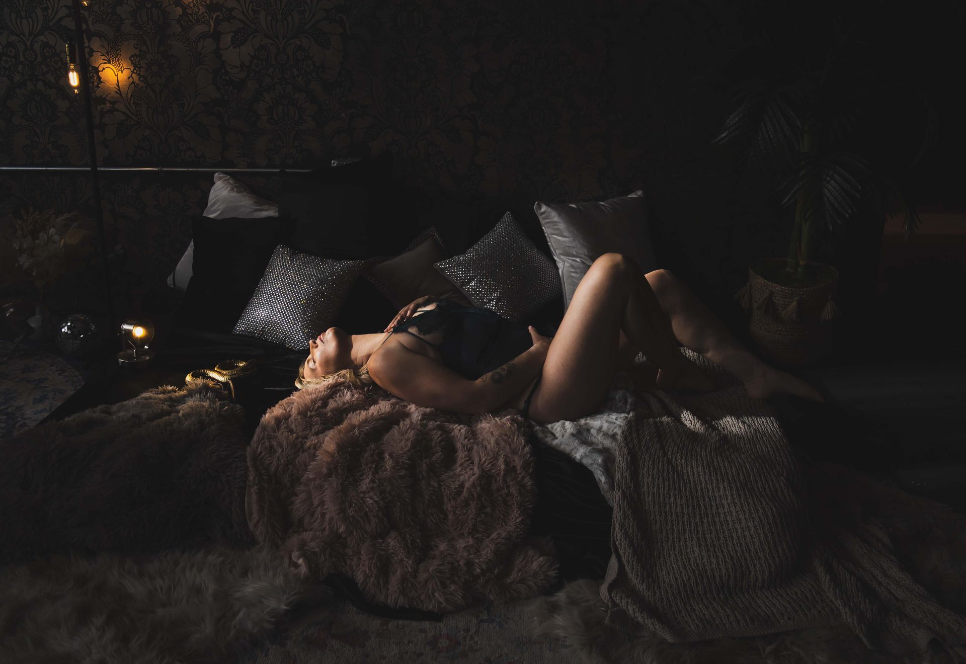 Woman reclines on a bed with soft pillows and blankets, dimly lit with fairy lights, creating a cozy atmosphere.