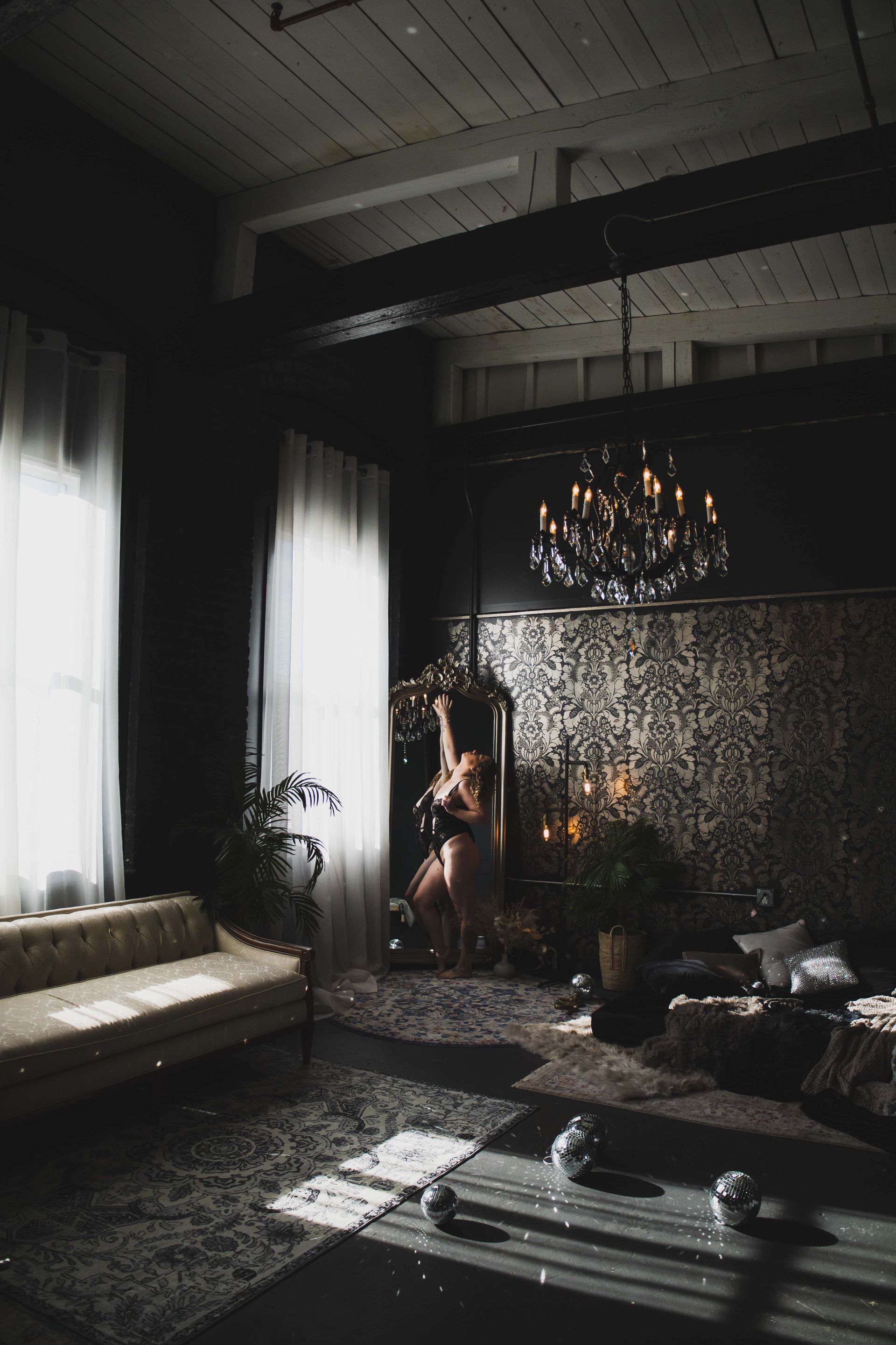 A woman in lingerie poses by a mirror in a dimly lit room with a chandelier and sofa. Sunlight streams in.