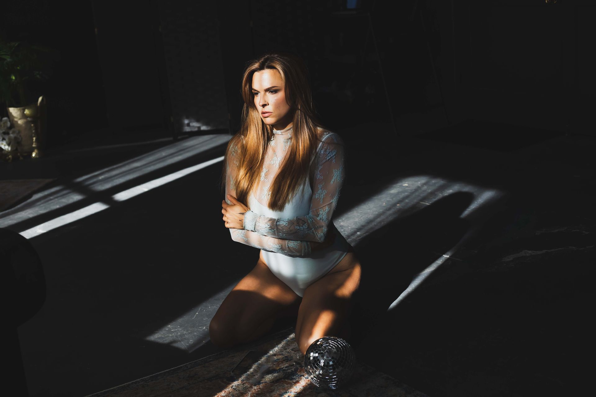 Woman in sparkly white bodysuit kneels, illuminated by sunlight. She looks off-camera, arms crossed, in a dark, indoor setting.