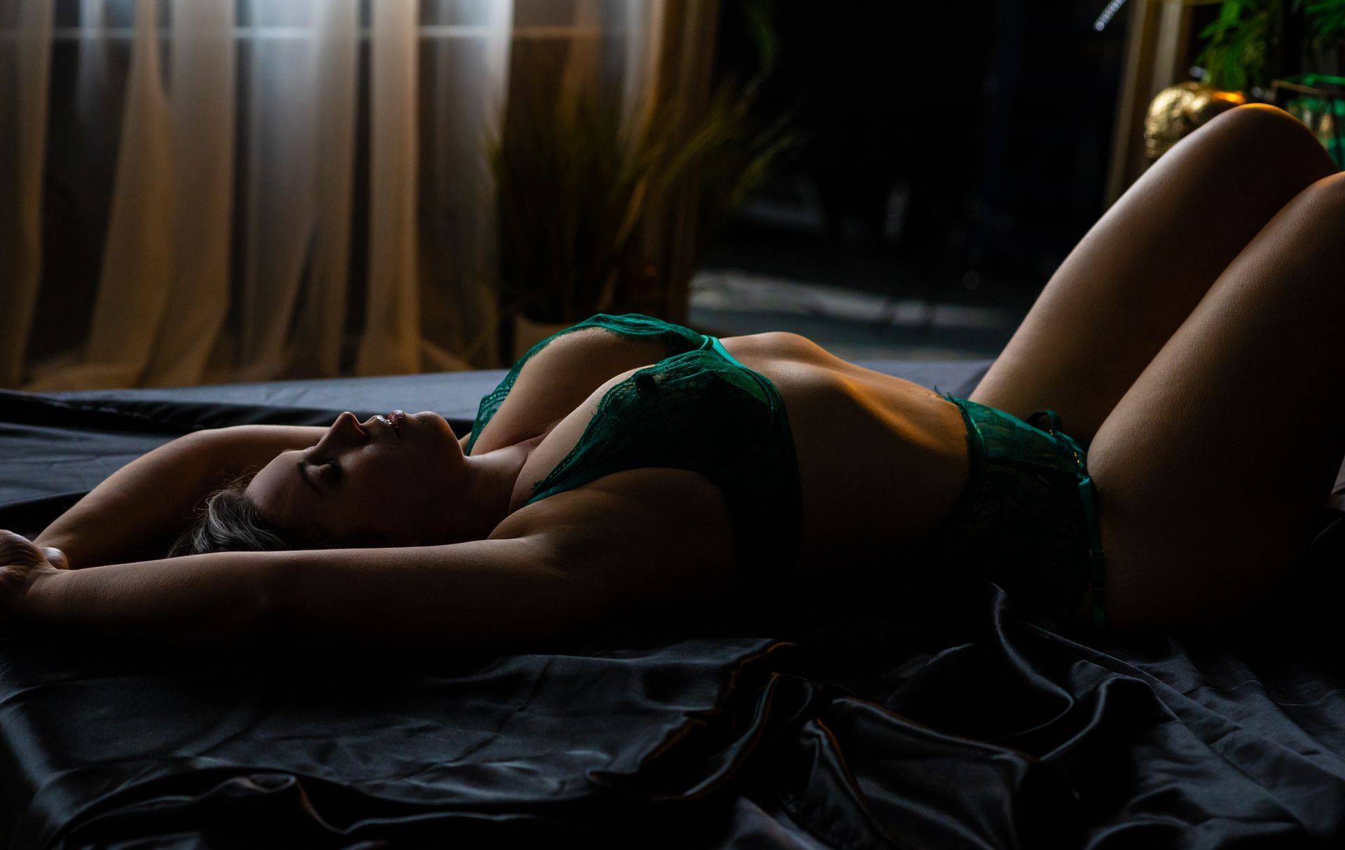Woman in green lingerie reclines on a bed with arms raised. Dim lighting with a dark setting.