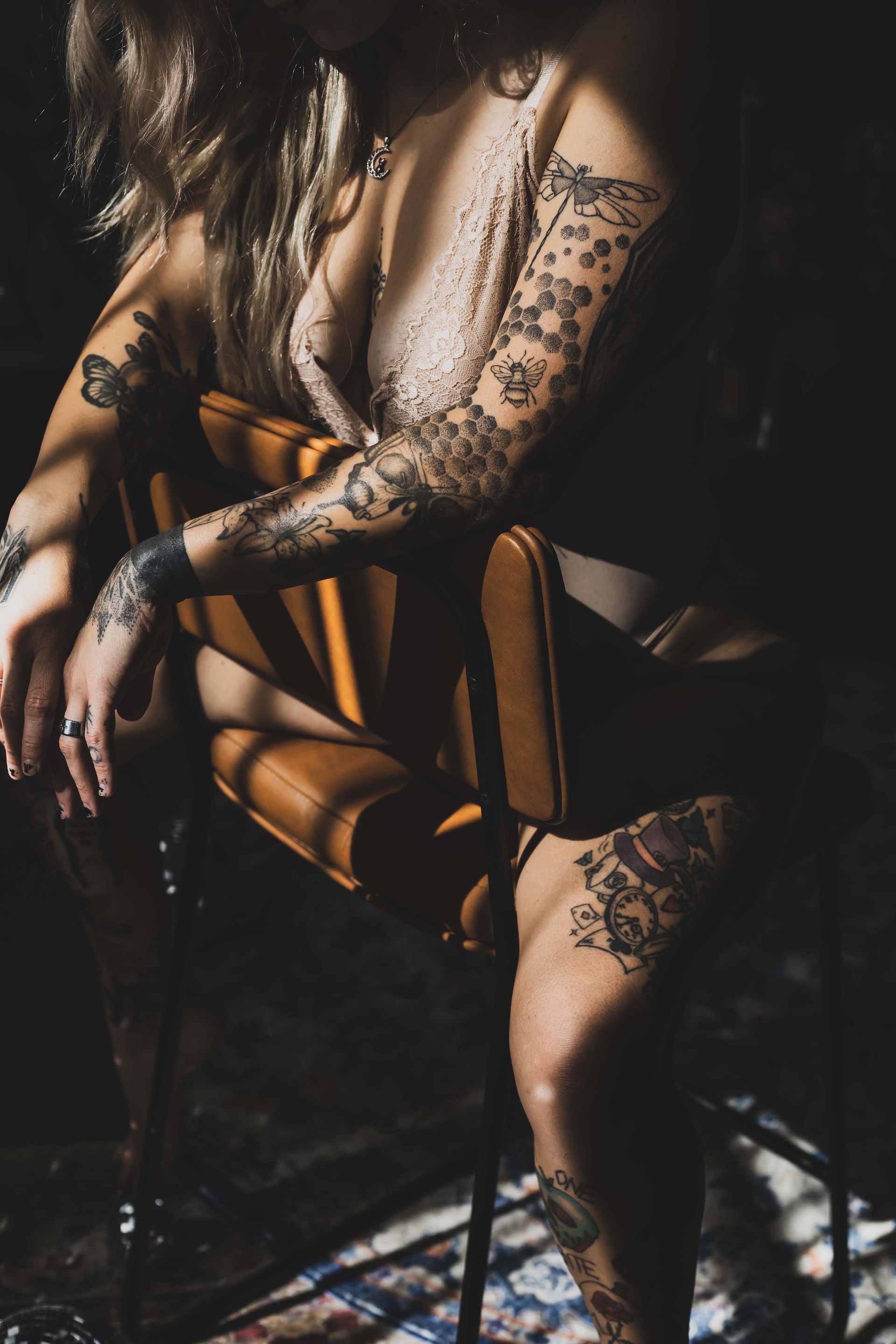 Woman with tattoos sits in a modern chair, lit by sunlight creating shadows. Dark setting.