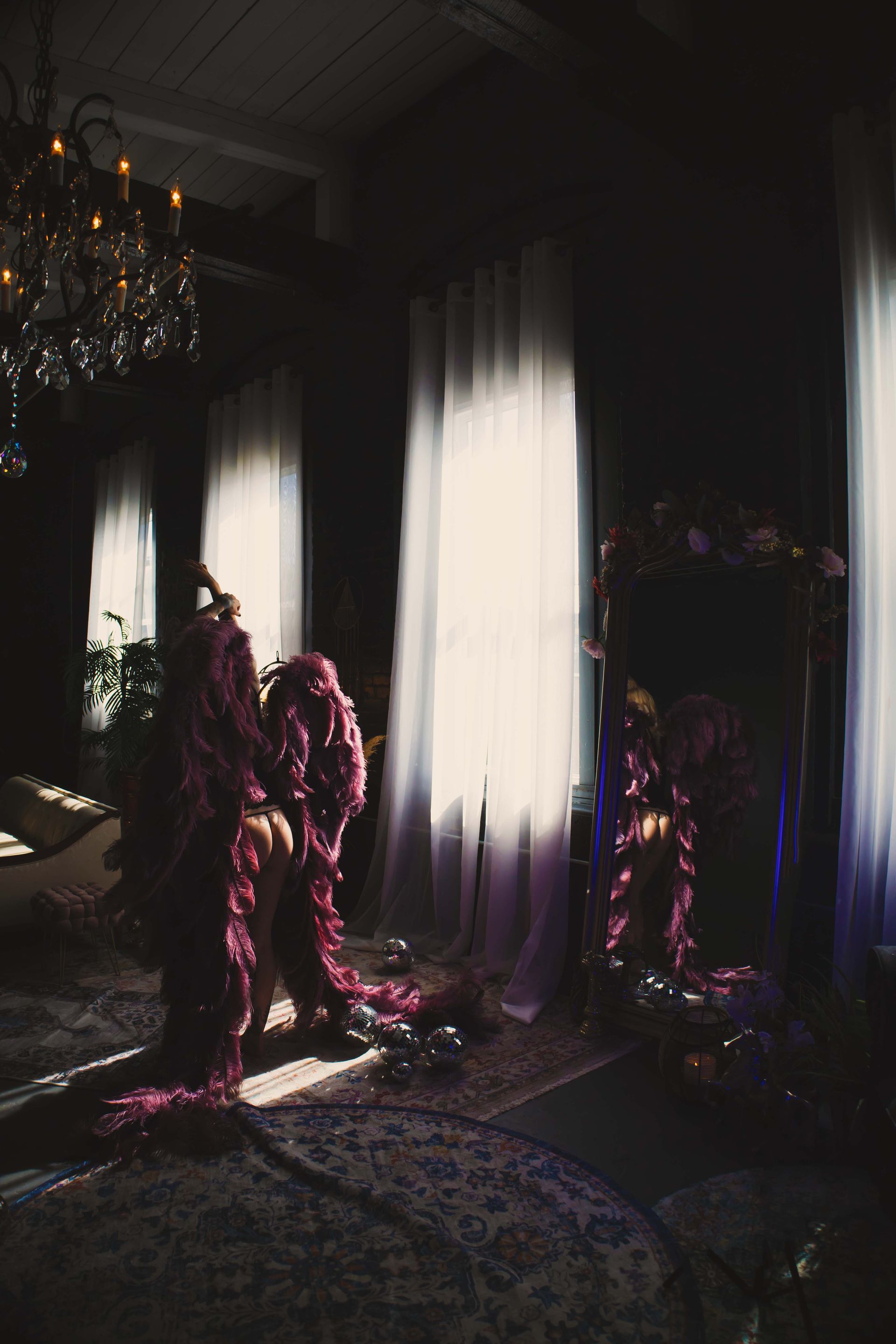 A darkly lit room with a figure draped in red fabric, standing near a mirror and a window with bright light. A chandelier hangs from the ceiling.