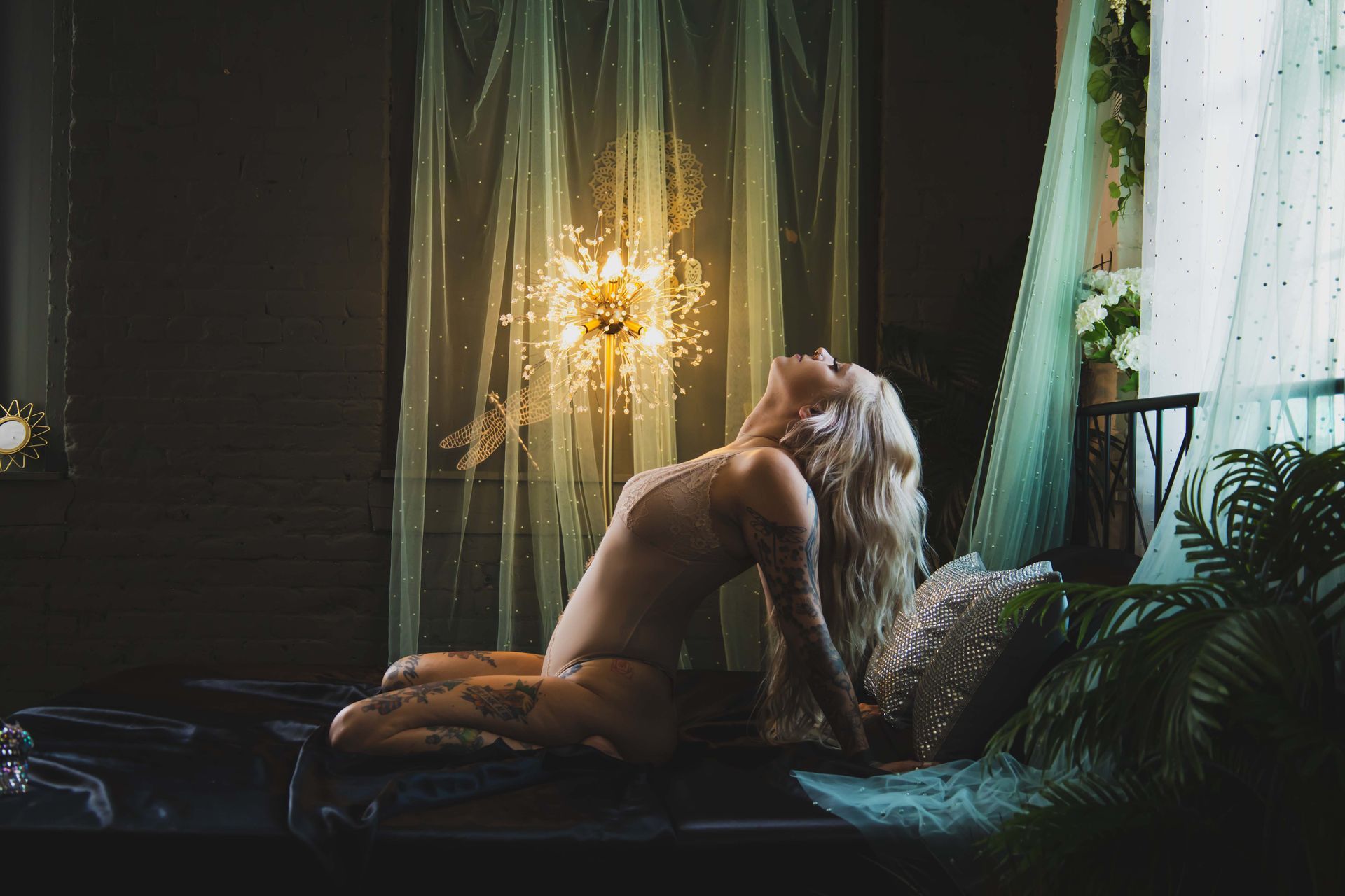 A blonde woman with tattoos reclines on a bed, looking upwards towards a glittering light fixture. Dark room with draped fabric and plants.