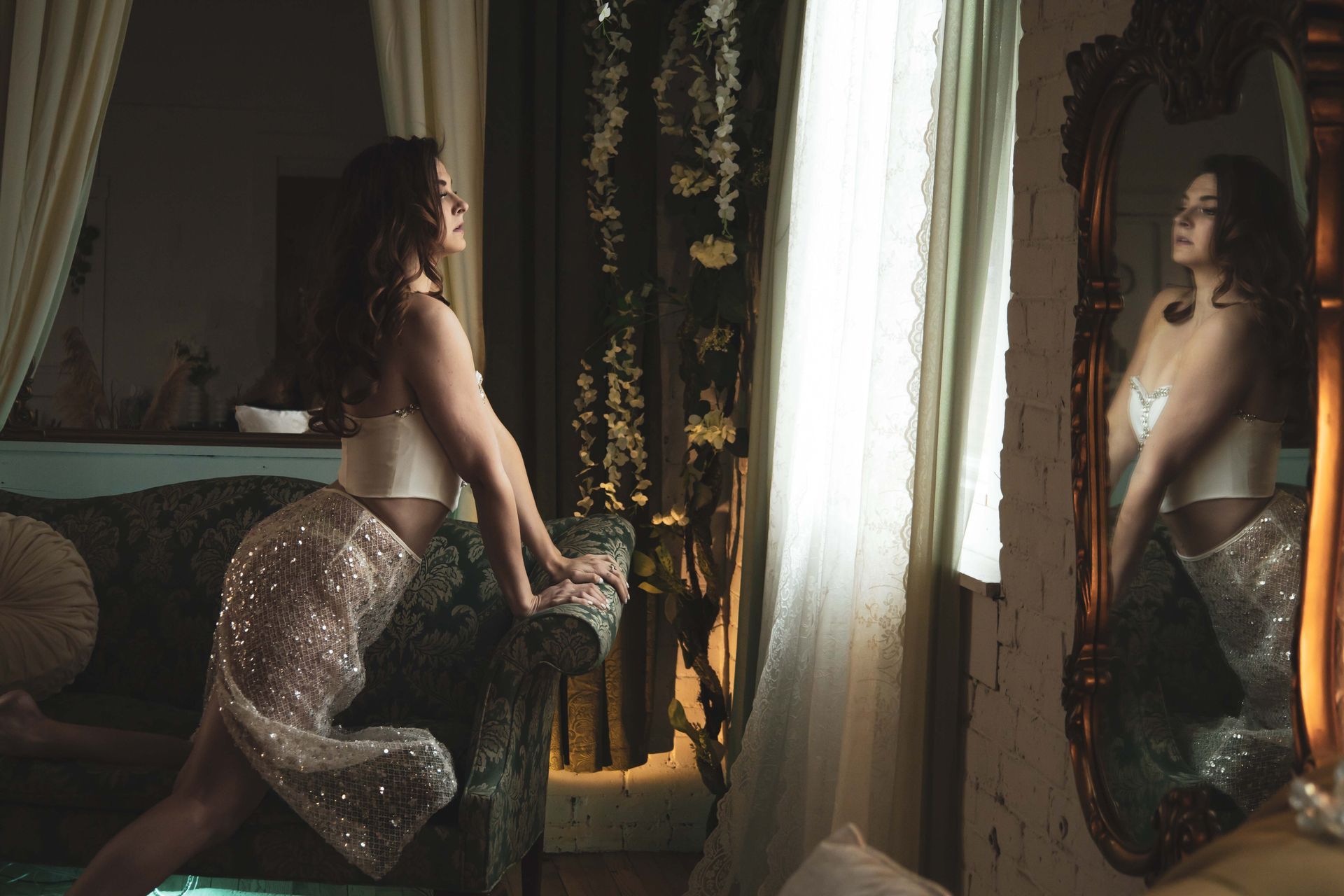 Woman in sparkling skirt and top leans against a green sofa, gazing at her reflection in a large antique mirror, near a window.