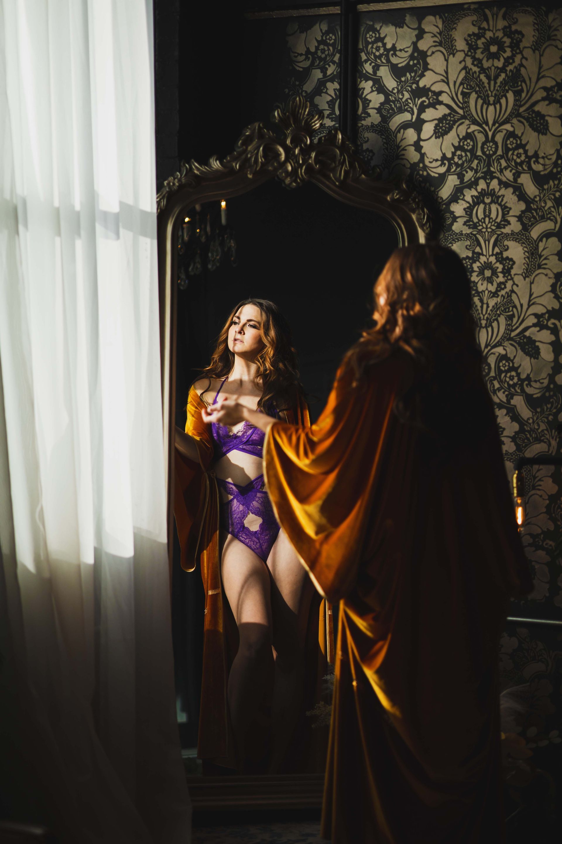 Woman in lingerie and robe looking in a decorative mirror. Sunlight streams in from a nearby window illuminating the scene with warm light.